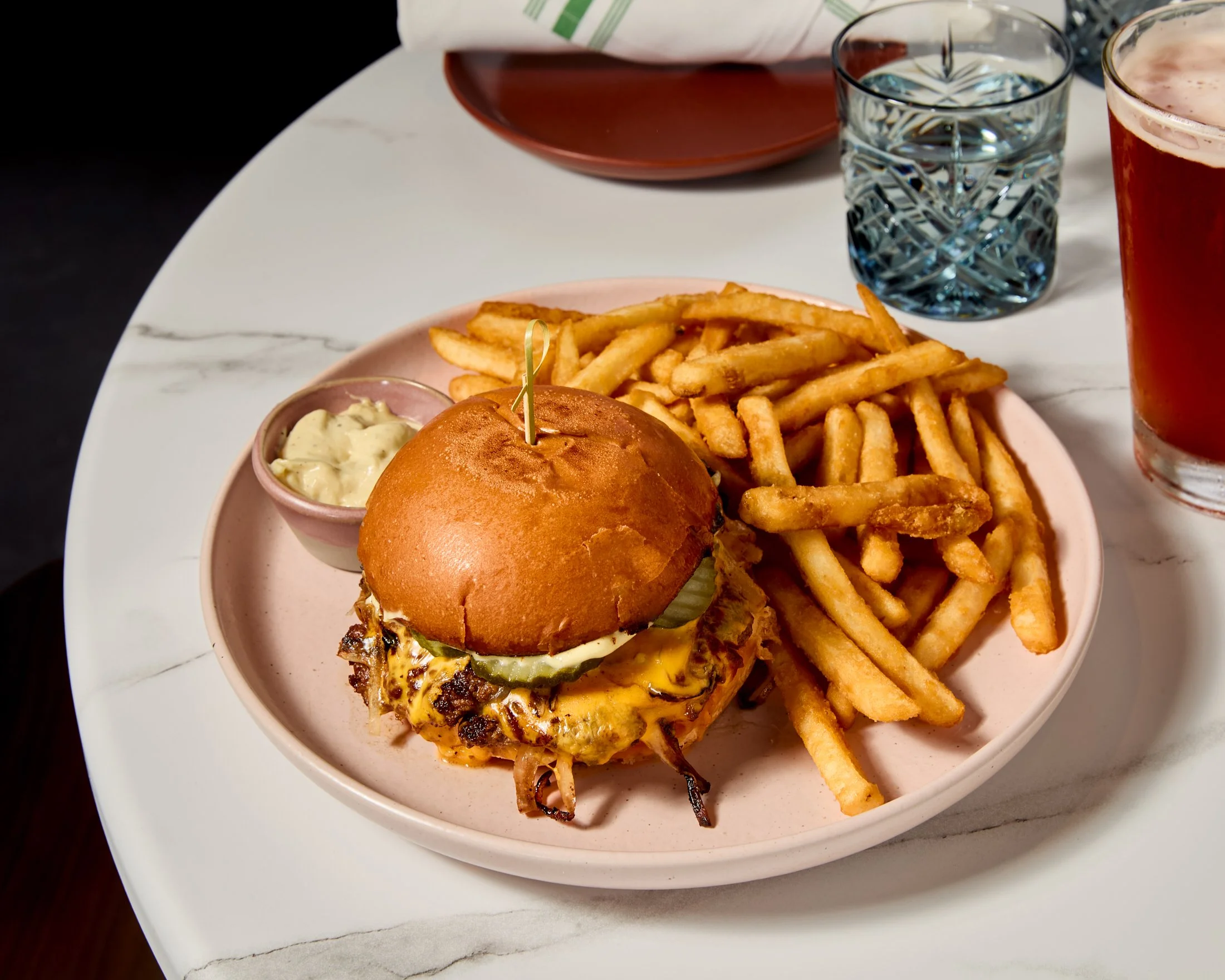 A plate with French fries, a burger with cheese, pickles, bacon, and a bun, served with a dipping sauce, along with two glasses of water and a glass of pink beverage, on a white marble table.