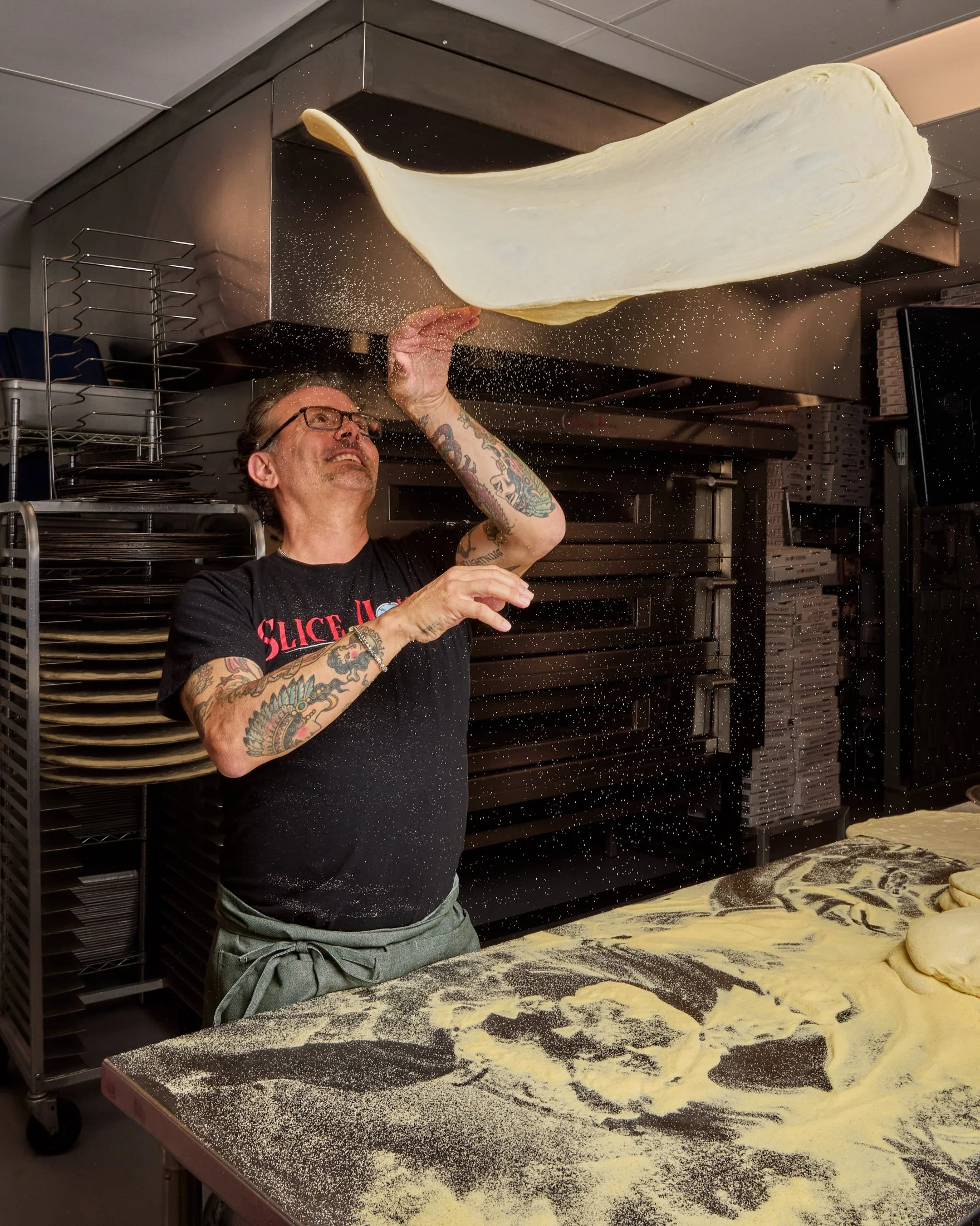 A man with tattoos and glasses is tossing pizza dough into the air in a commercial kitchen. Flour is scattered on a countertop with rolled-out dough pieces, and shelves with pizza trays and boxes are visible in the background.