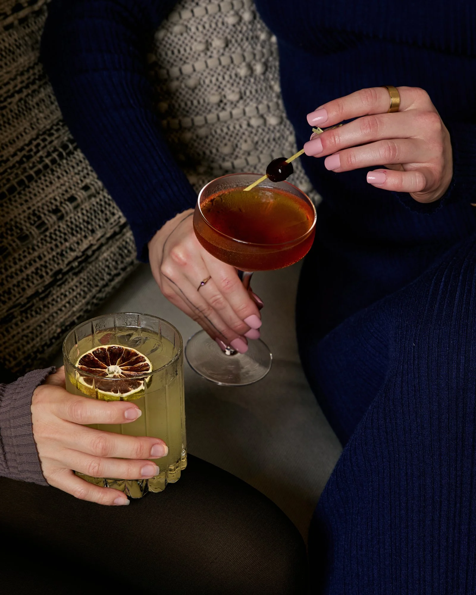 Two people holding cocktails, one with a lemon slice and the other with a cherry garnish.