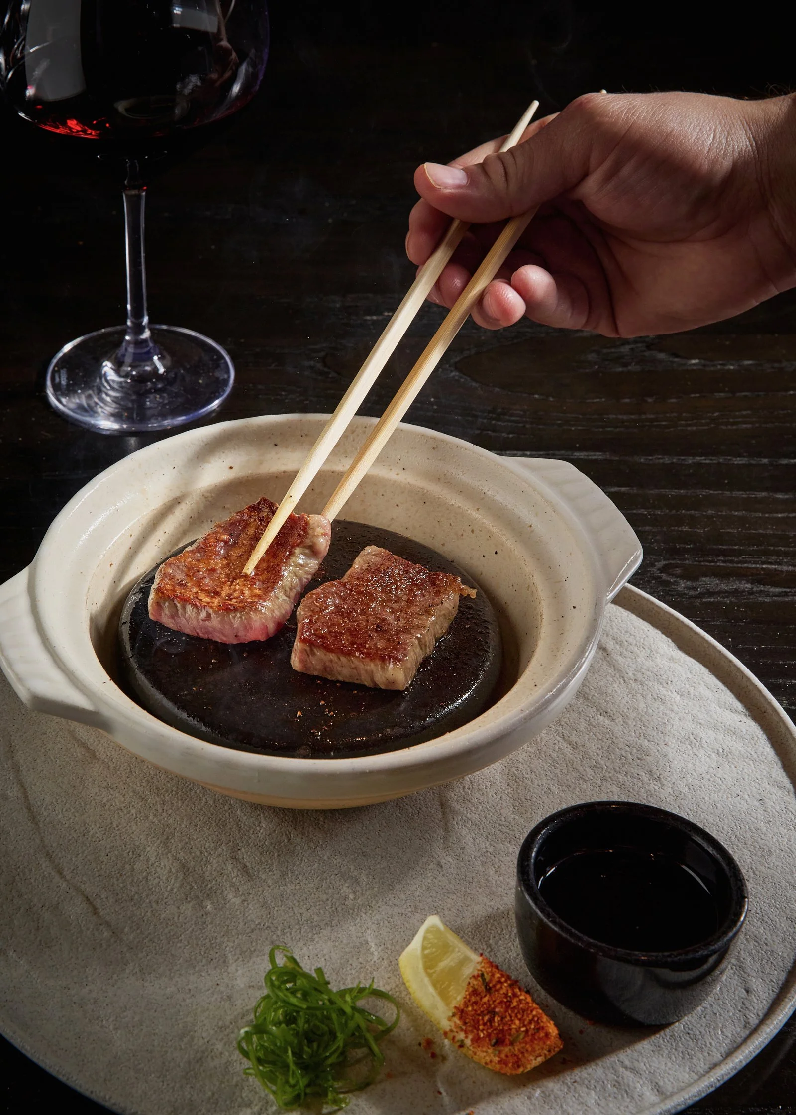 A hand using chopsticks to pick up a piece of grilled steak from a small black grill dish, with a glass of red wine and a dipping sauce on a beige plate.