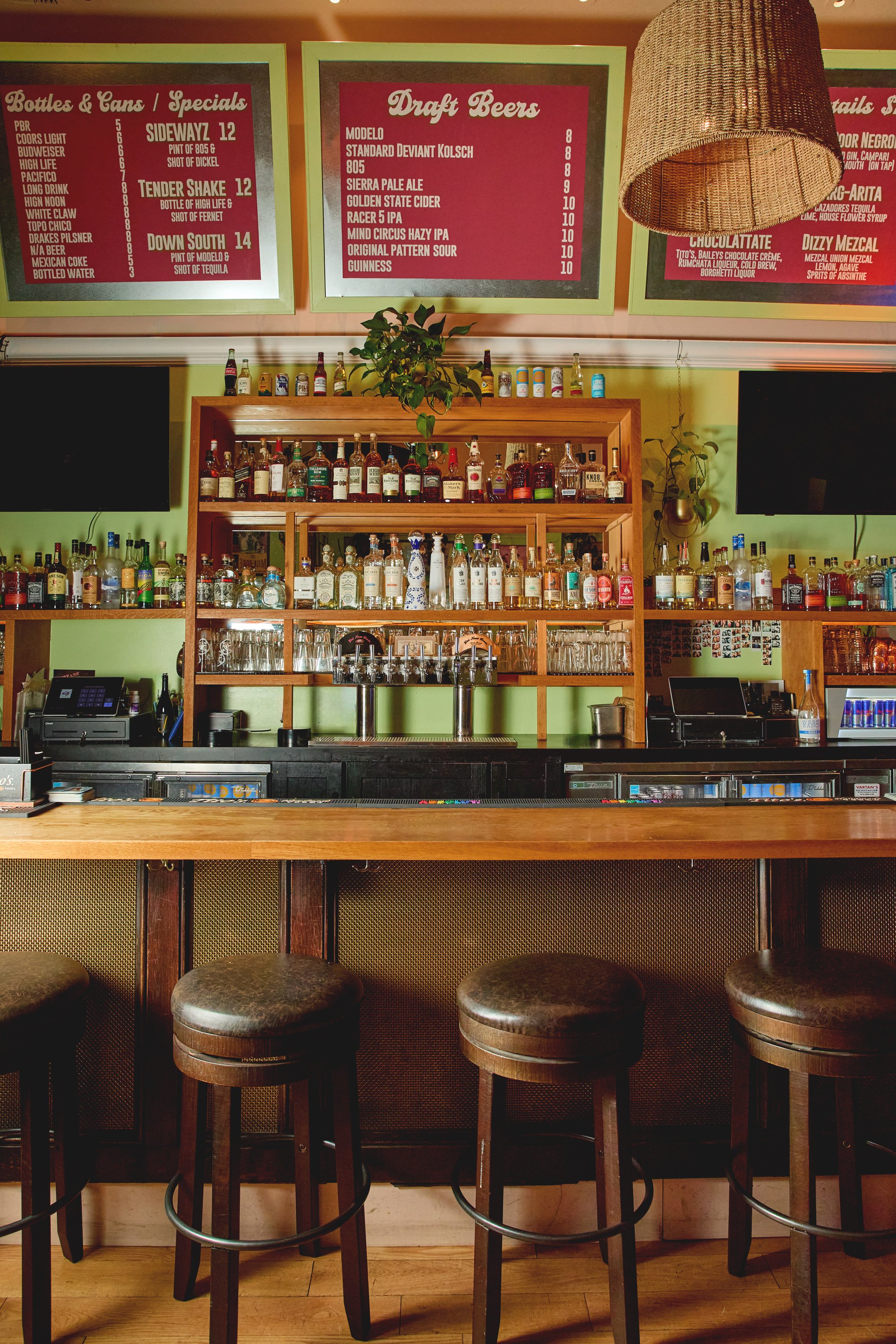 A bar with a wooden counter and four bar stools in the foreground. Behind the counter, shelves display various bottles of alcohol. Above, two large green-framed menus list draft beers and drink specials. A plant is on top of the shelves, and a woven 