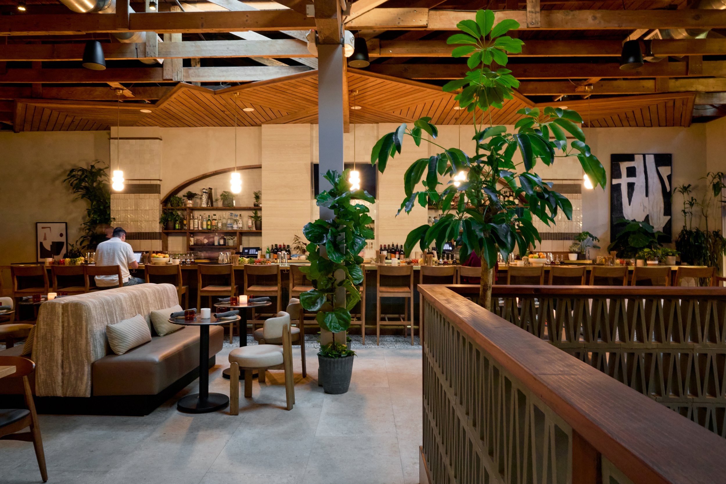 Interior of a restaurant with wooden accents, a bar area in the background, a person sitting at the bar, green plants, and a cozy seating area with beige and gray furniture.