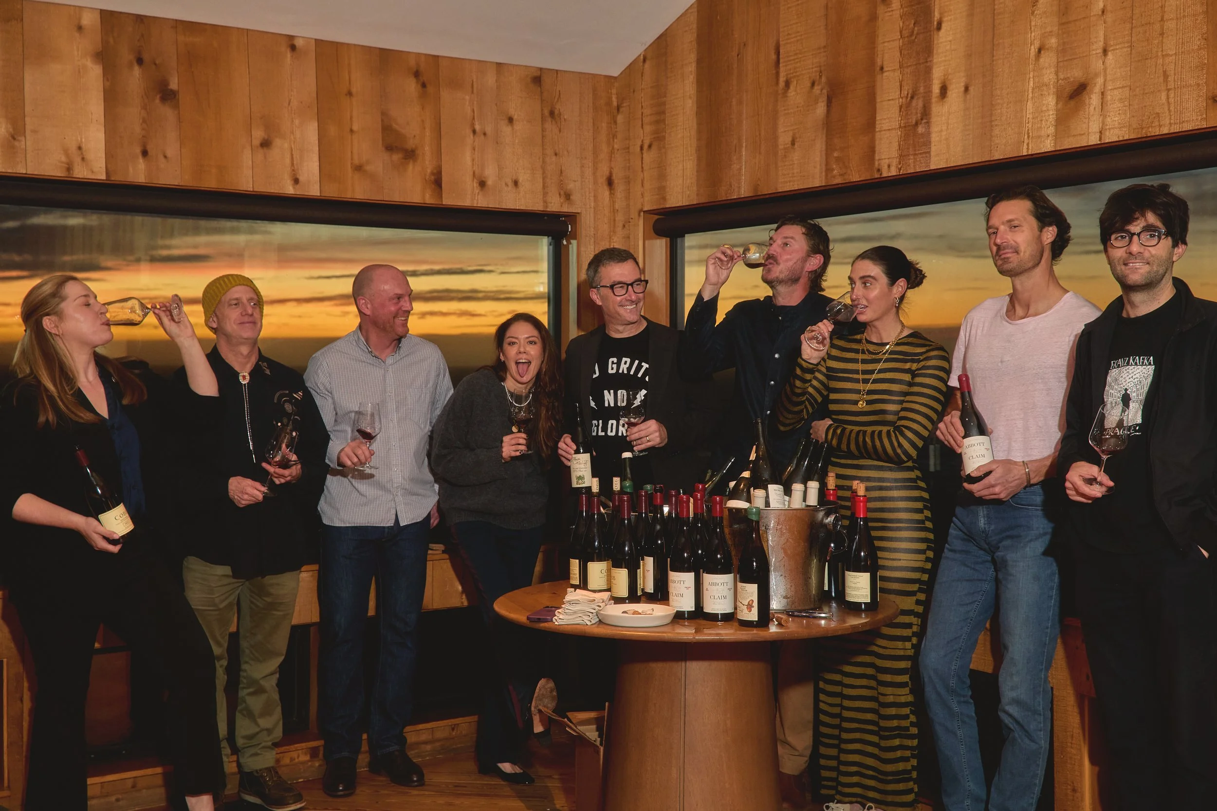 Group of people celebrating with wine bottles at a wooden-paneled room with sunset views outside windows.