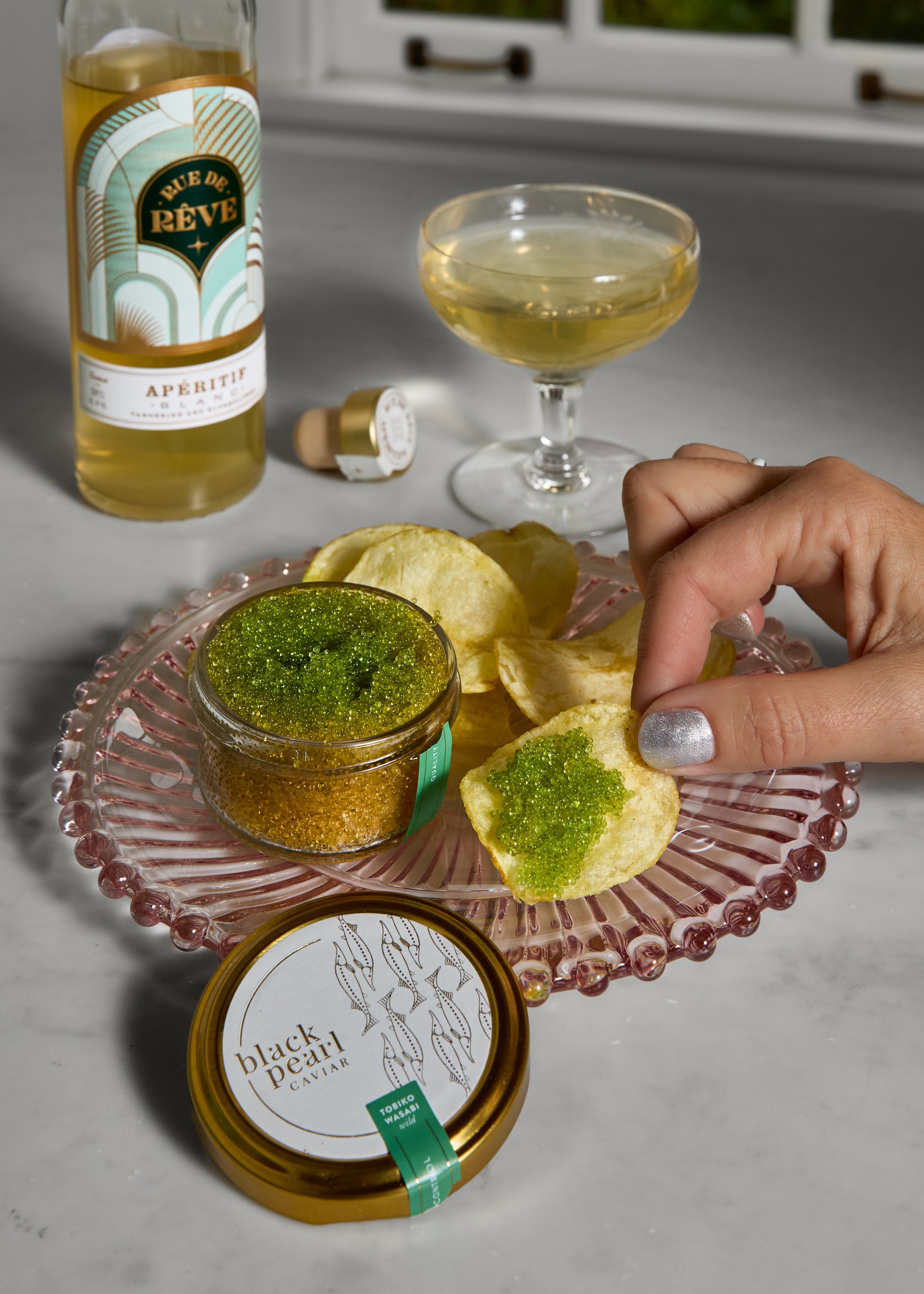A pink glass plate holds potato chips and a small jar of green and gold caviar, with a hand reaching to pick up a chip. In the background, a glass of white wine, a bottle of apéritif, and a container of caviar are visible on a gray surface.