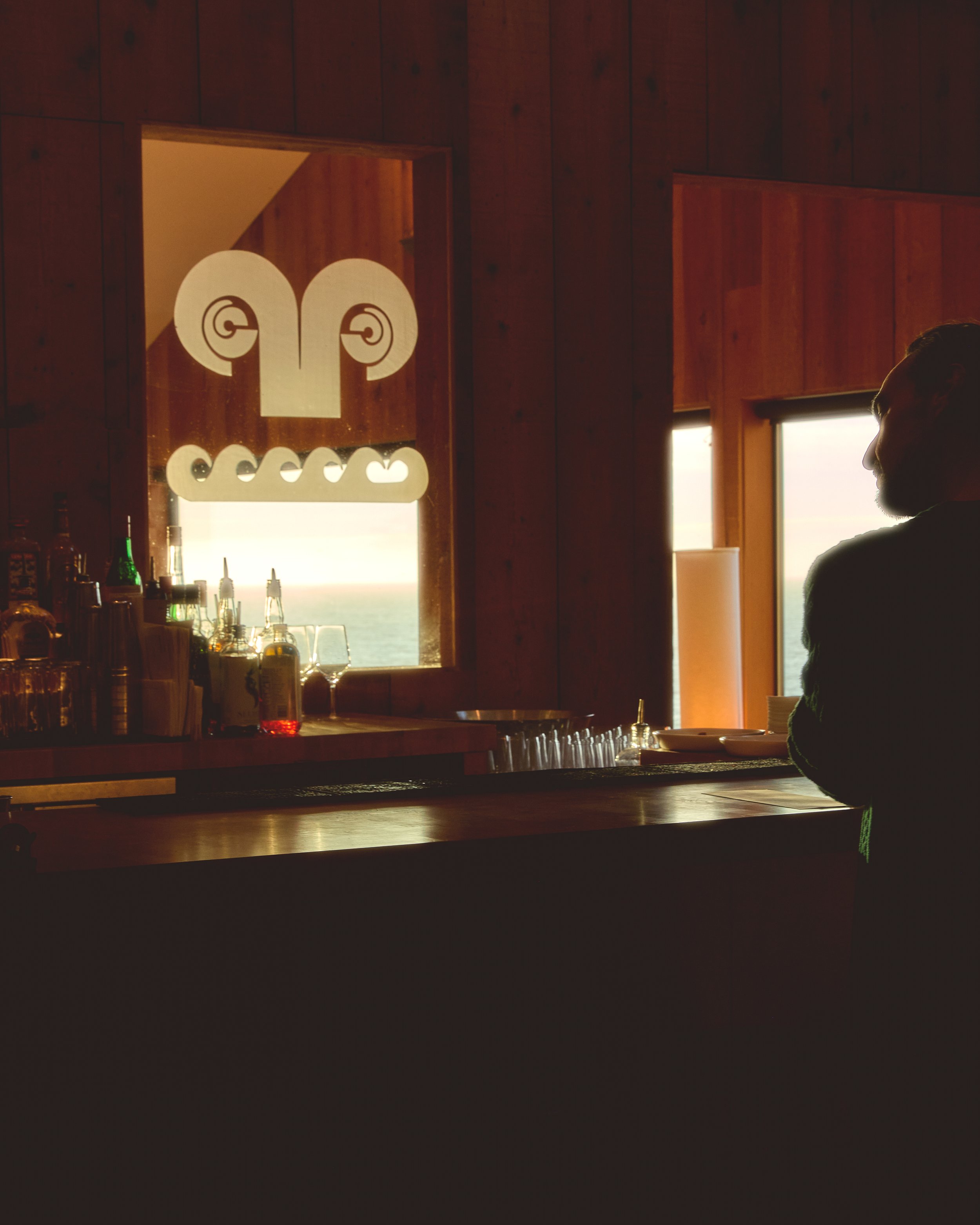 Silhouette of a man standing at a bar with wooden paneling, ocean view through windows, and tribal art window decoration.