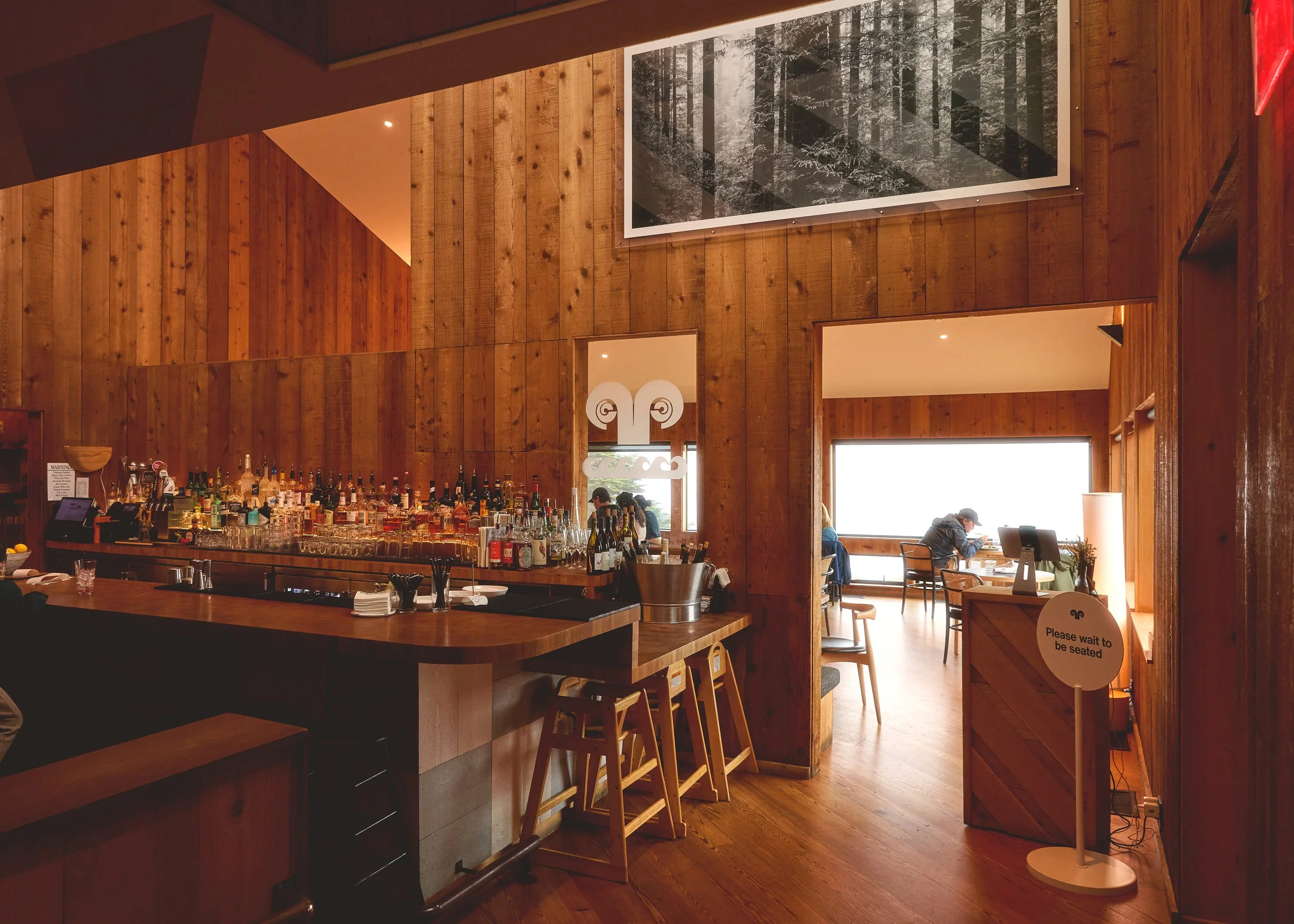 Interior of a cozy restaurant with a wooden bar, filled with bottles of liquor, and a sign that says "Please wait to be seated." There are diners sitting at tables near large windows flooded with natural light.