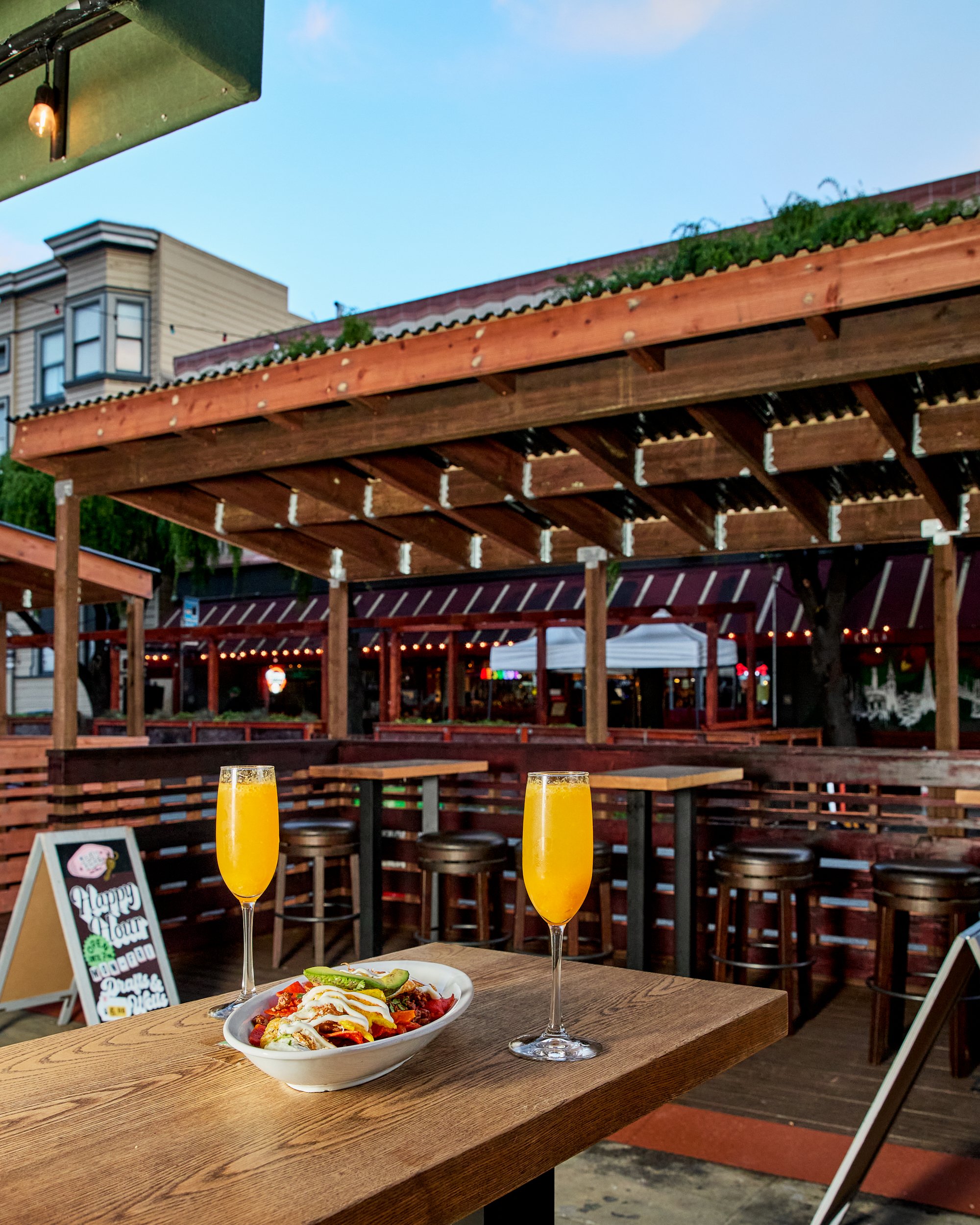 An outdoor patio at a restaurant or bar with a wooden table holding a bowl of food and two glasses of orange drink. The background has a wooden pergola and a sign that says 'Happy Hour'.