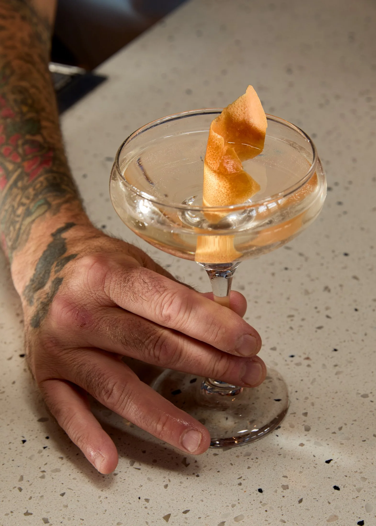 A person with tattoos on their arm holding a cocktail glass with a lemon peel garnish on a speckled countertop.