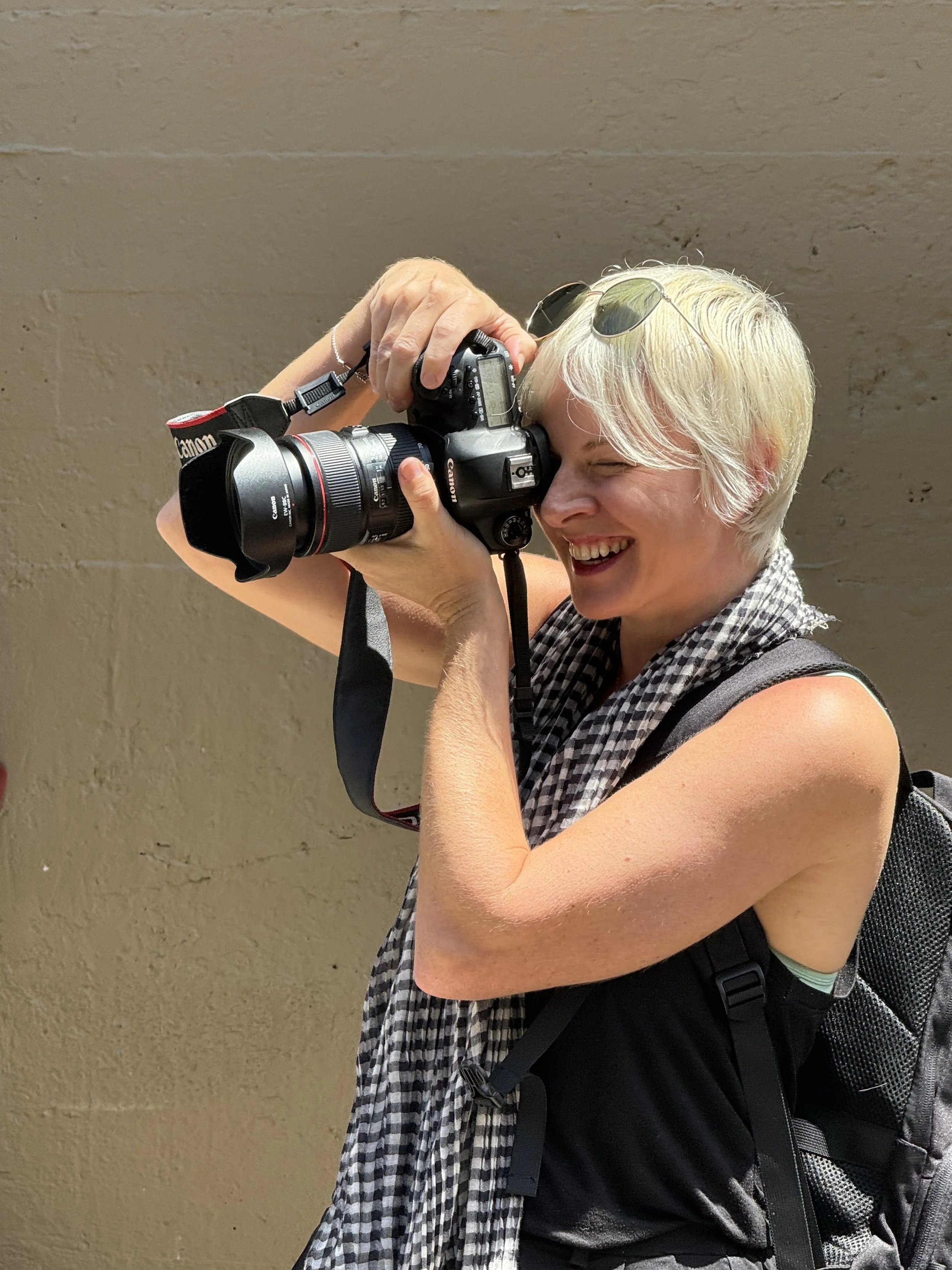 A woman with blonde hair smiling as she takes a photograph with a Canon camera, standing against a plain beige wall, wearing sunglasses on her head, a black shirt, and a carbon backpack.