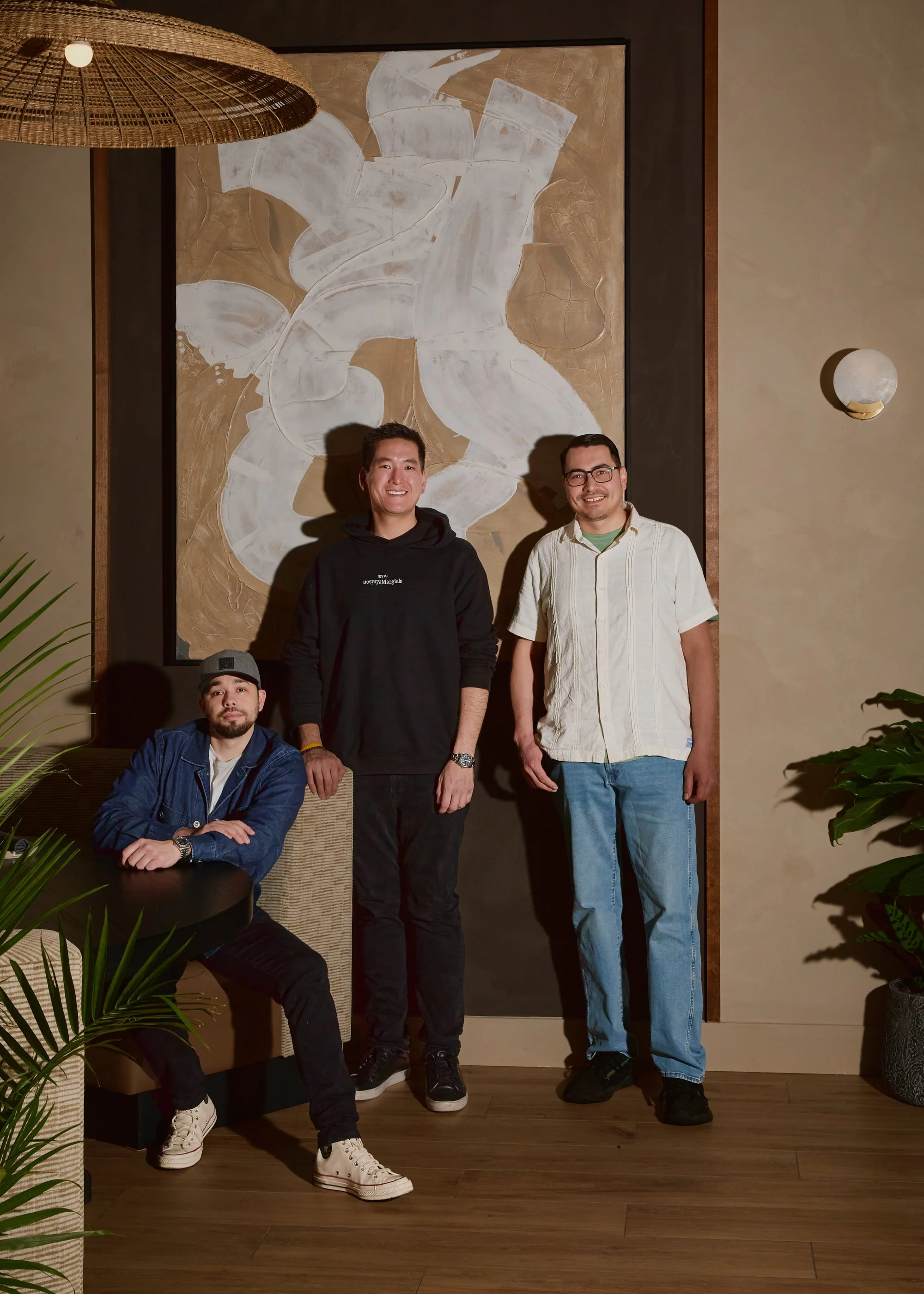Three men in casual clothing standing and sitting in front of a large abstract painting in a modern interior space with plants and wooden flooring.