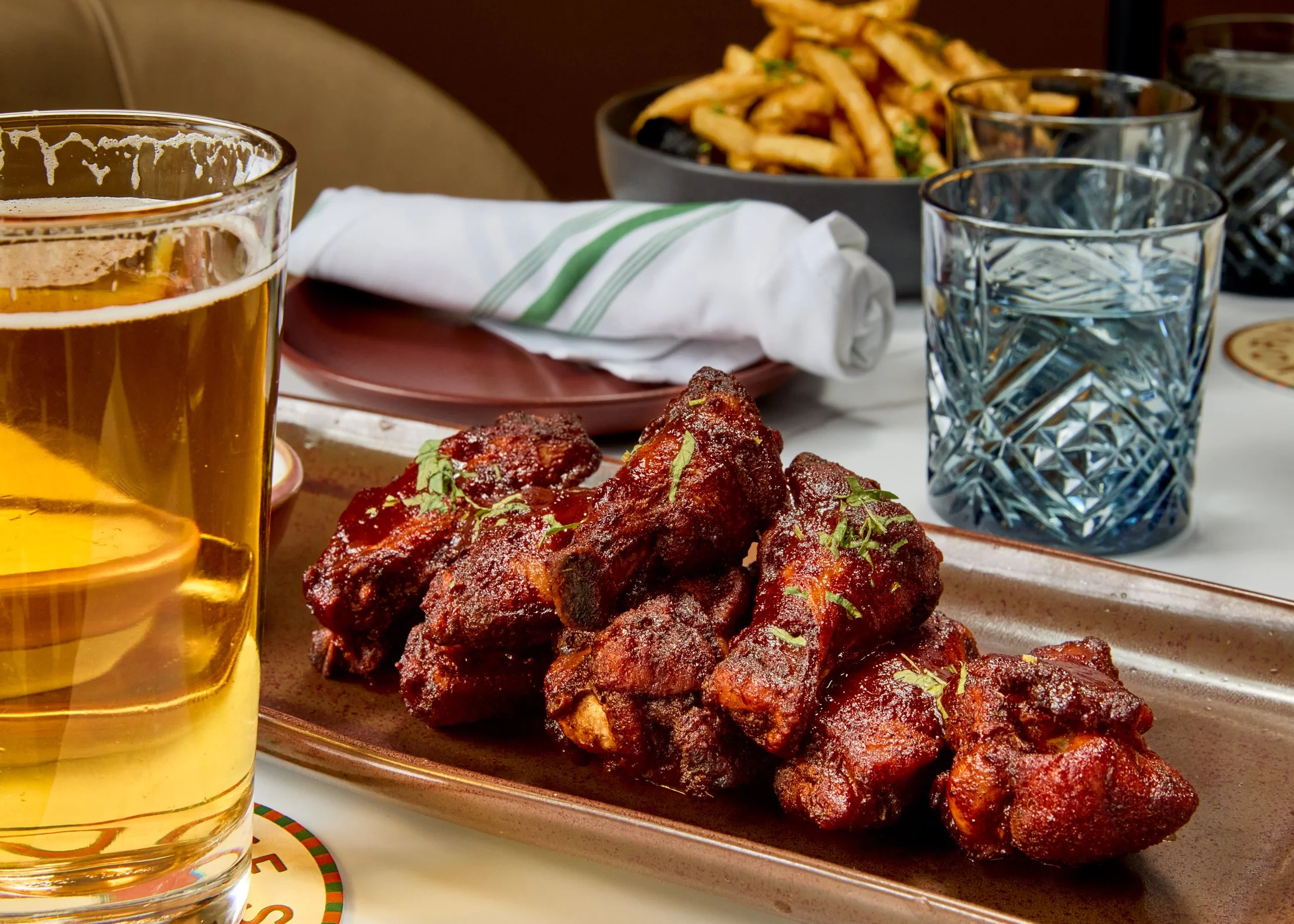 A glass of beer, a plate of spicy barbecue chicken wings garnished with herbs, a bowl of French fries with herbs, a drinking glass of water, some napkins, and utensils on a table.