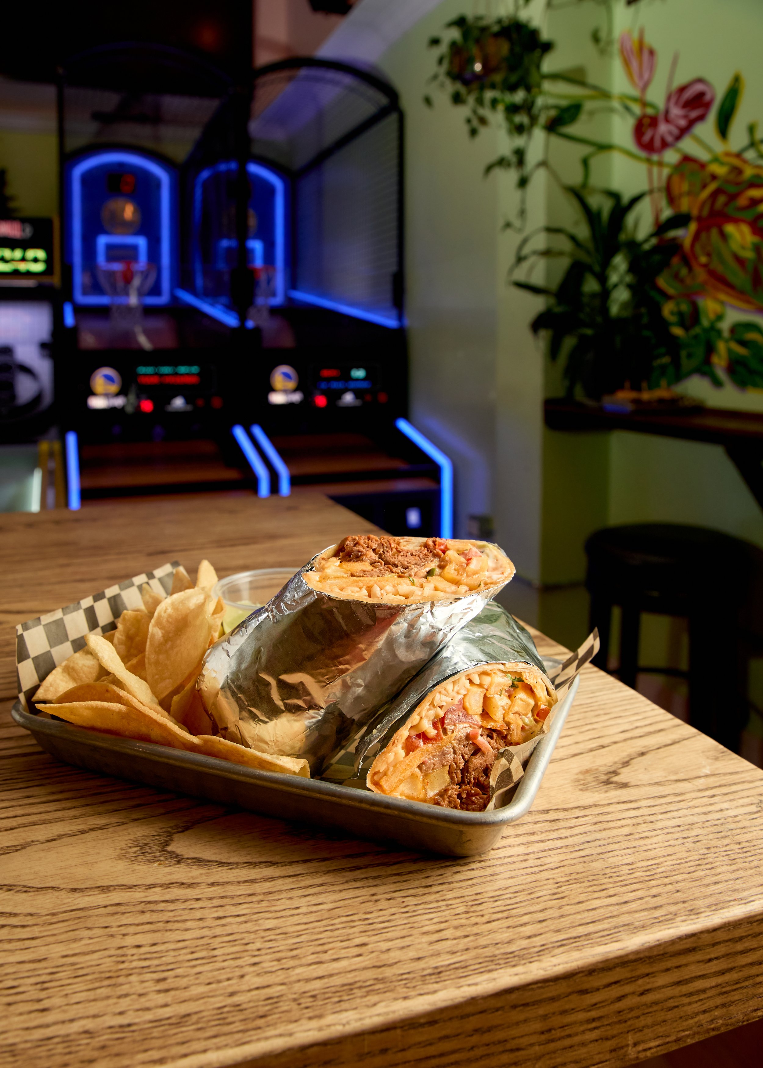 Two toasted wrap sandwiches filled with meat, cheese, and vegetables, served with potato chips in a metal tray on a wooden table at an arcade bar.
