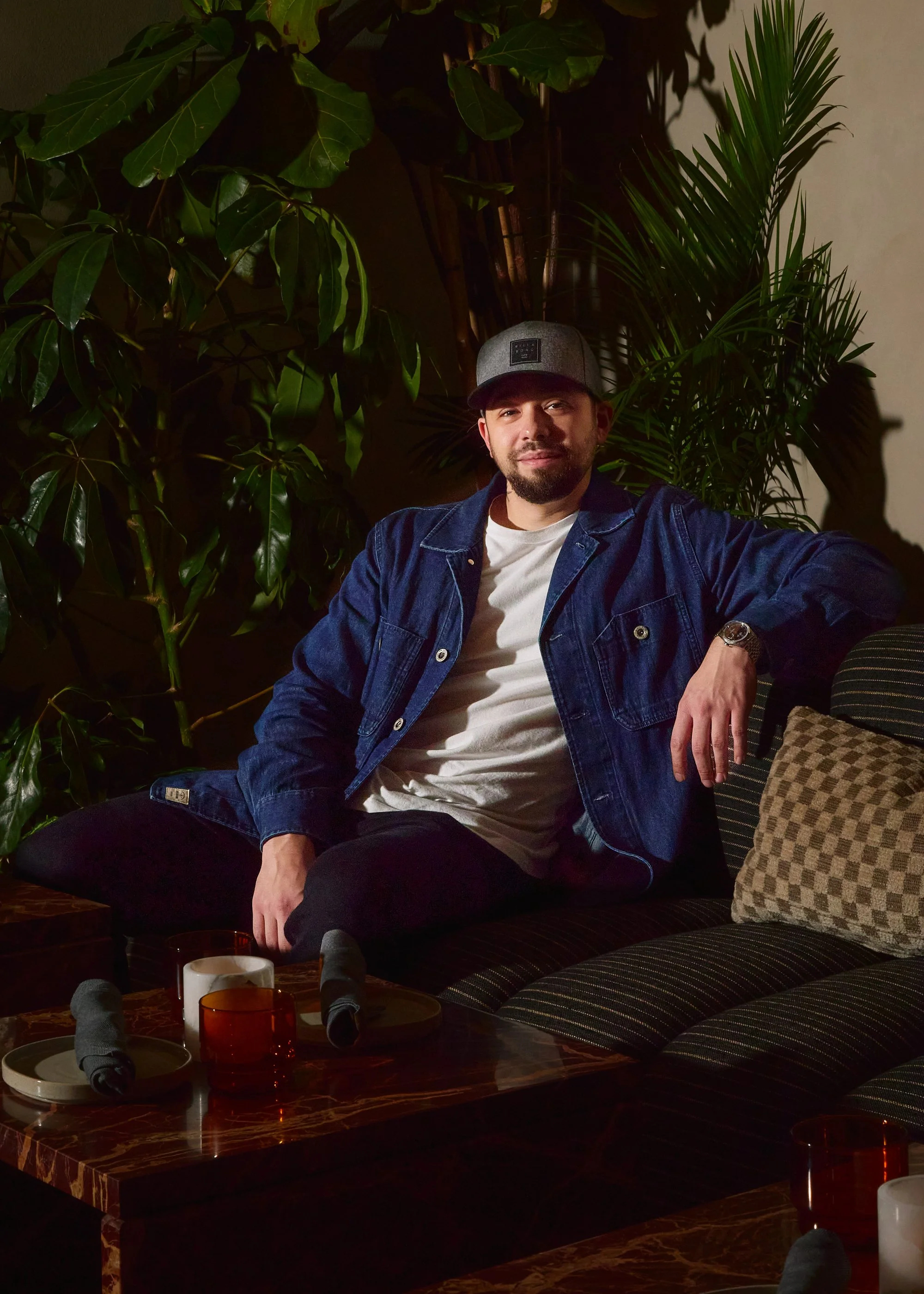 A man with a beard and mustache wearing a gray baseball cap, a white T-shirt, and a dark blue denim jacket, sitting on a patterned sofa in a dimly lit room with large green indoor plants in the background.