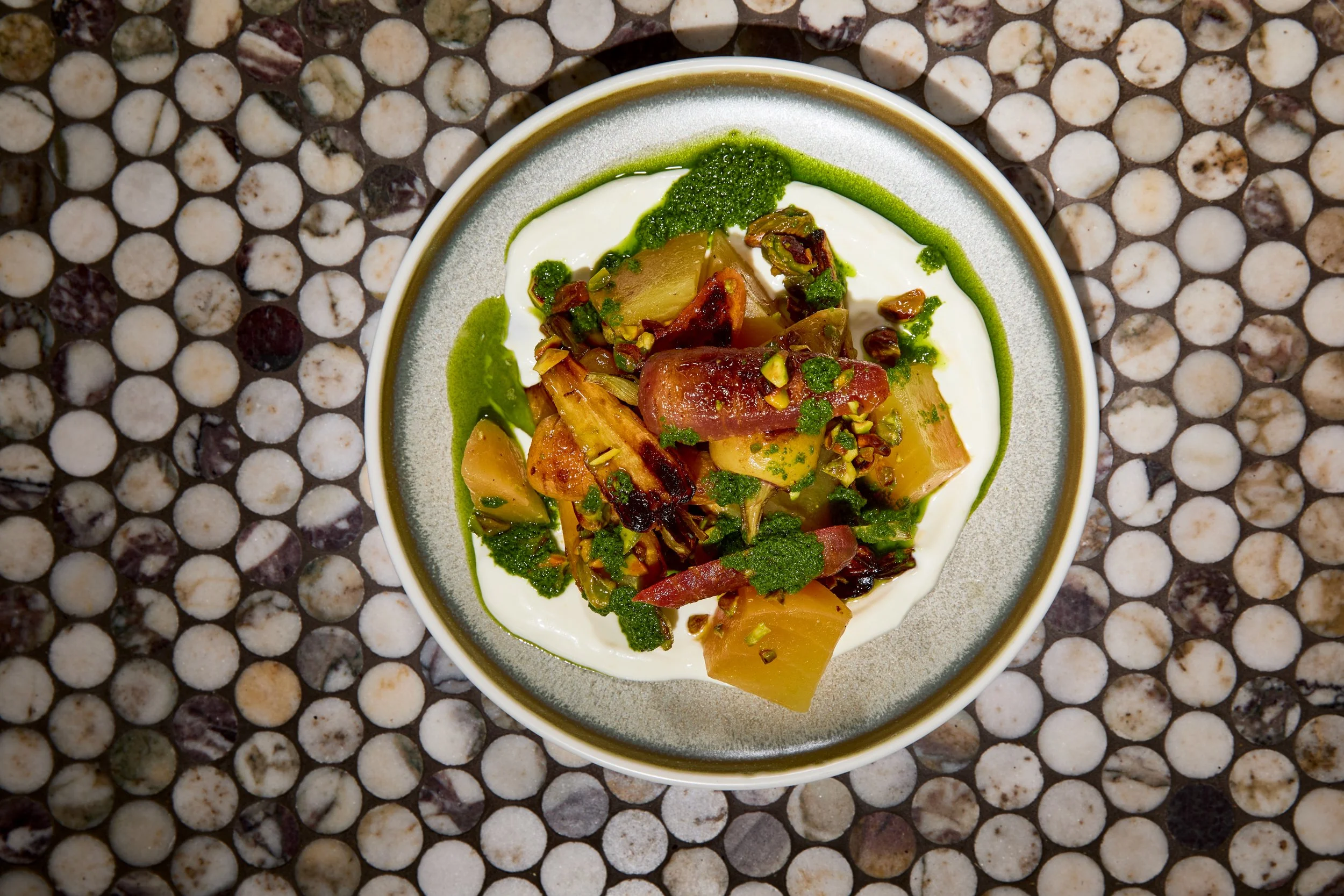 A bowl of yogurt topped with roasted vegetables, chopped pistachios, green sauce, and herbs on a mosaic tiled surface.