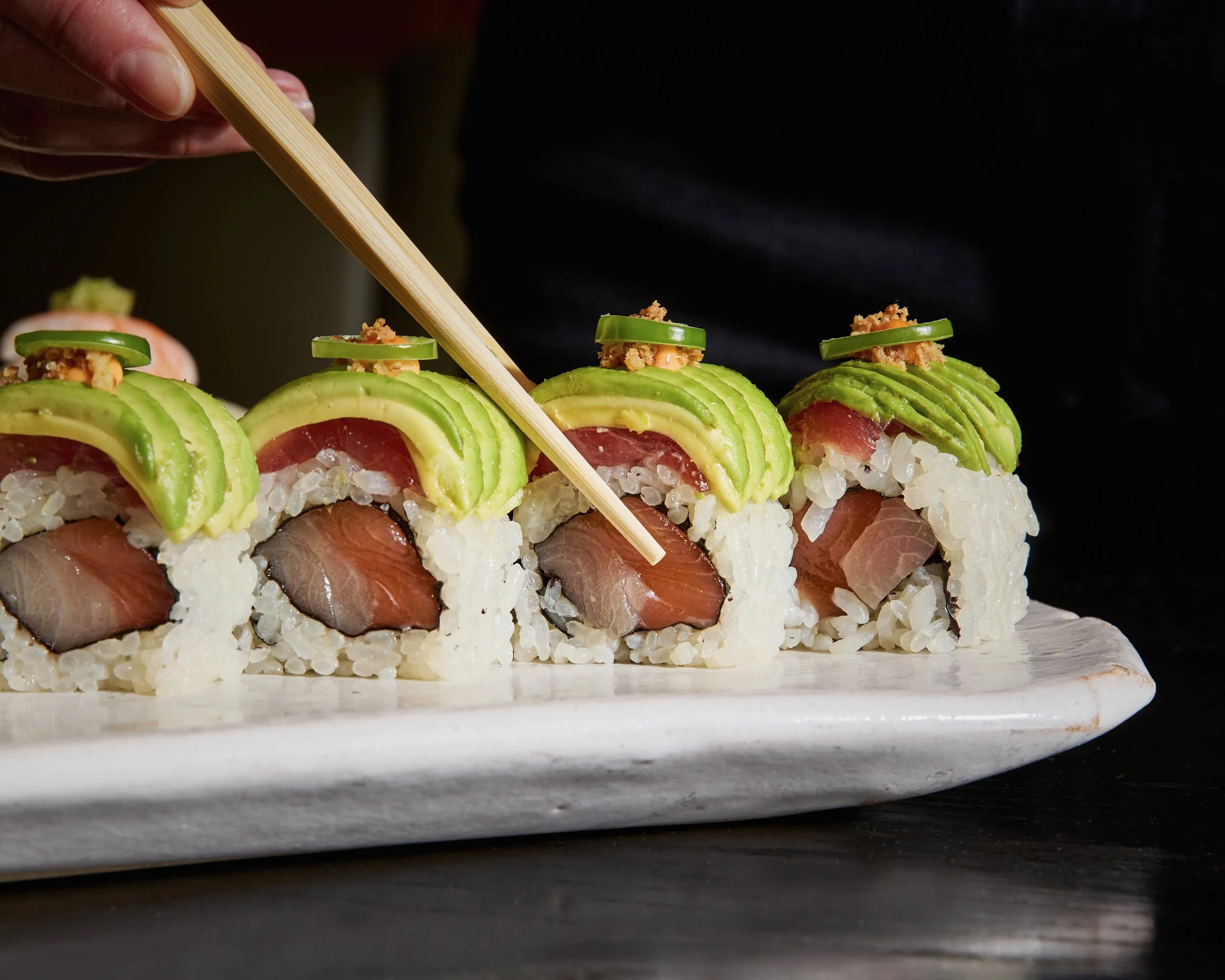 Close-up of sushi rolls topped with sliced avocado, cucumber, and fish, on a white platter with a black background.