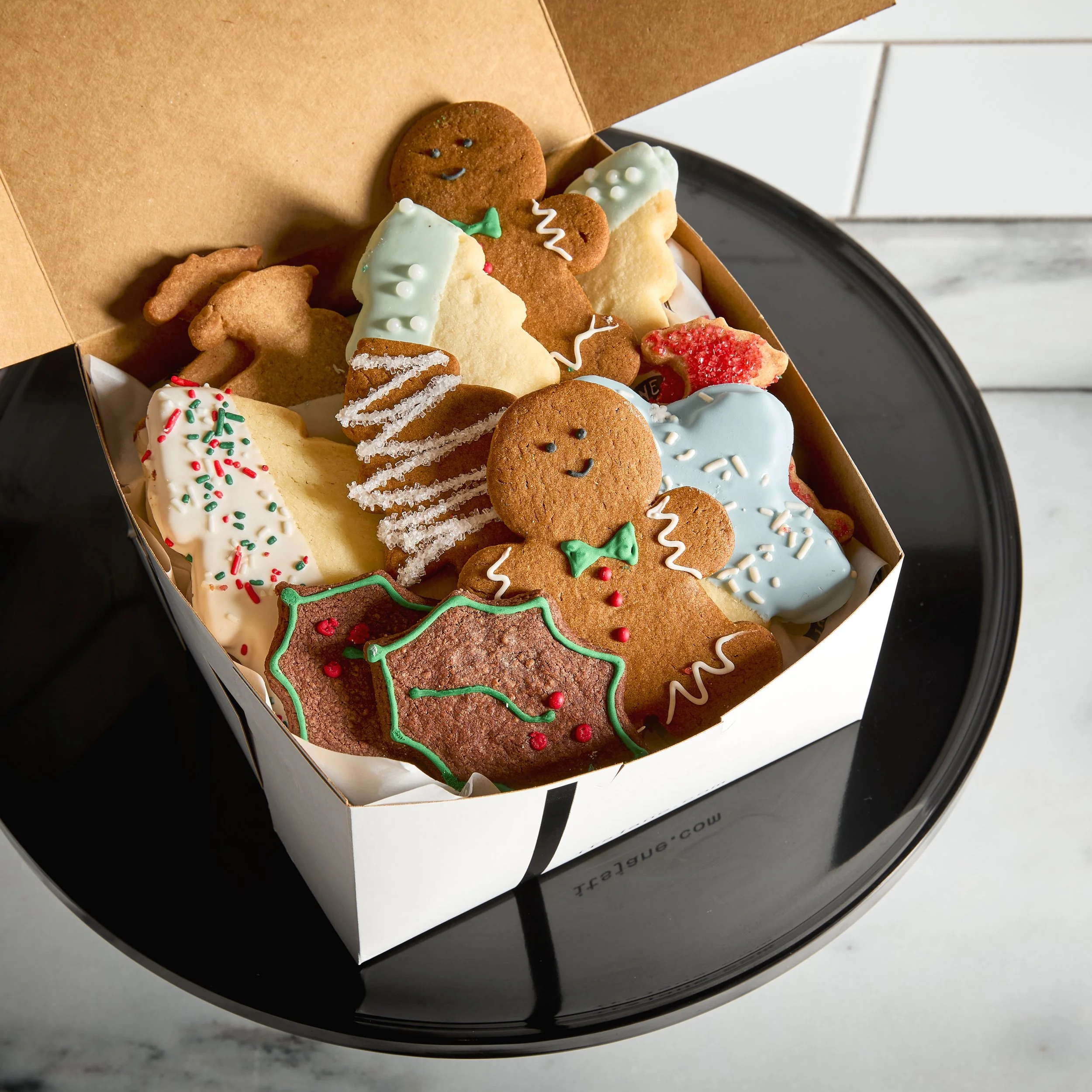 A box of decorated Christmas cookies on a black tray, featuring gingerbread men, holly-shaped cookie, sugar-coated cookie, and other festive treats with colorful icing and sprinkles.