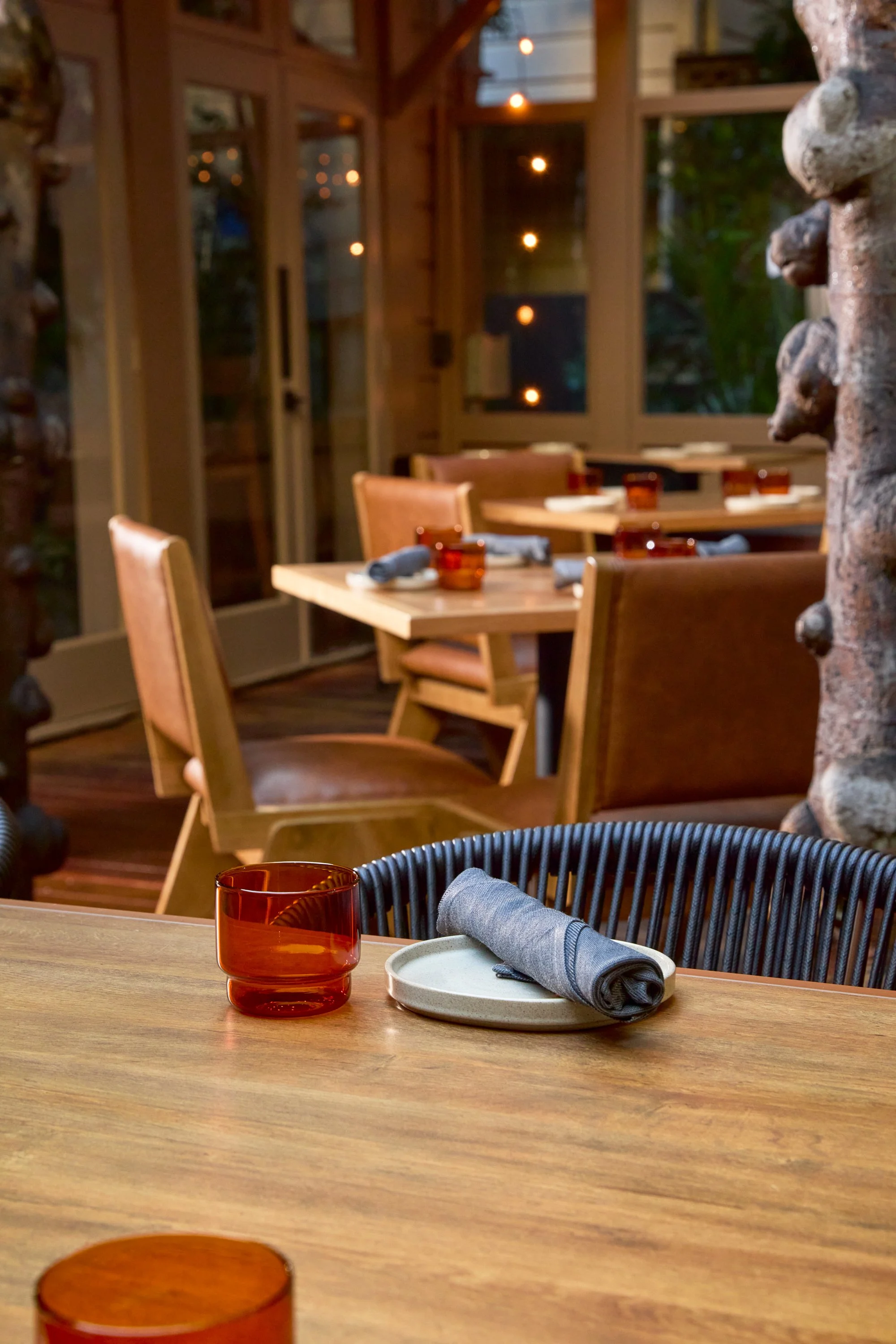 Close-up of a table set with a rolled gray napkin on a small beige plate and an amber glass, in a warmly lit restaurant interior with wooden furniture and multiple tables.