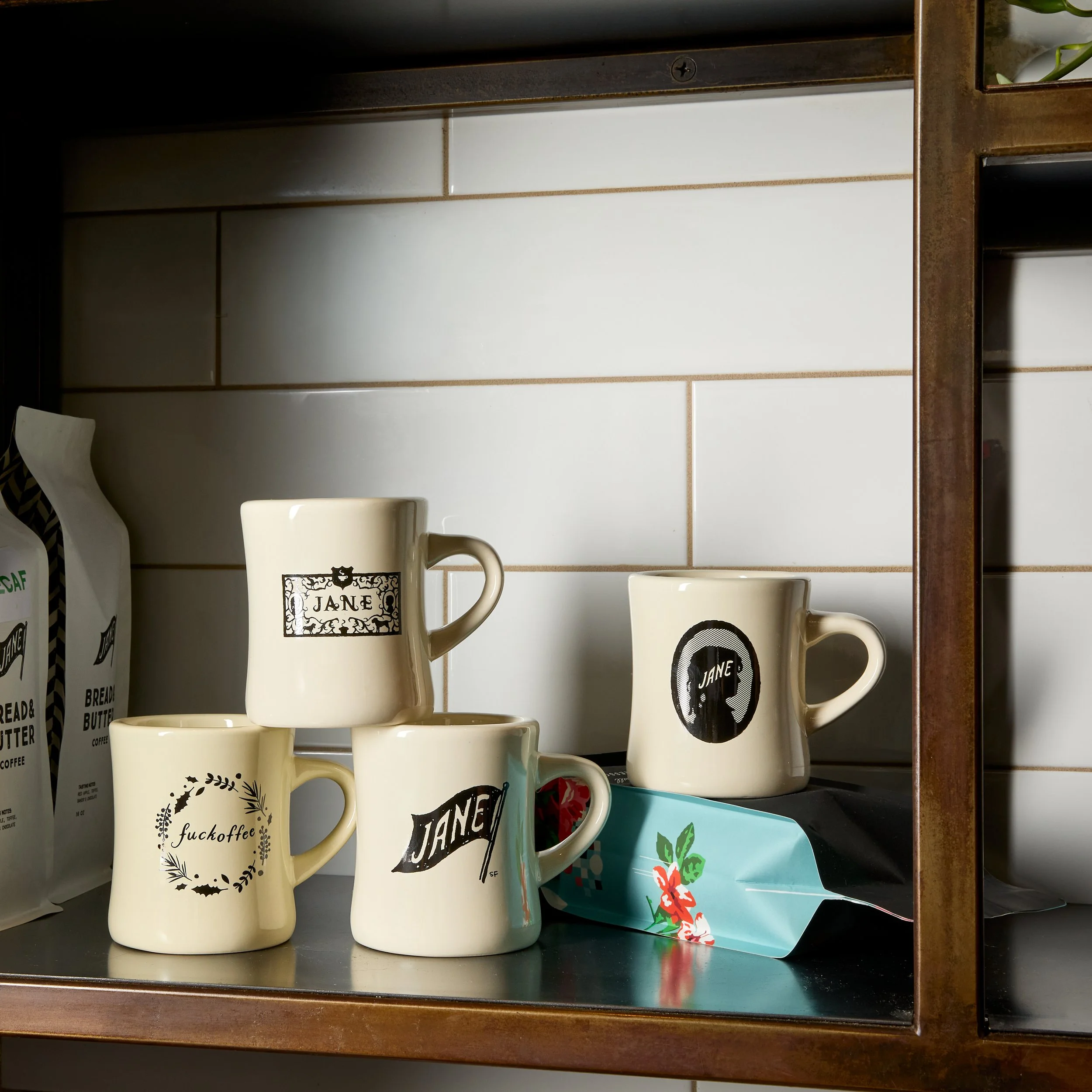 Collection of four coffee mugs with various black and white designs and text on a kitchen shelf with a brown wood frame, against a white tiled wall, and a blue tissue box with a floral pattern nearby.