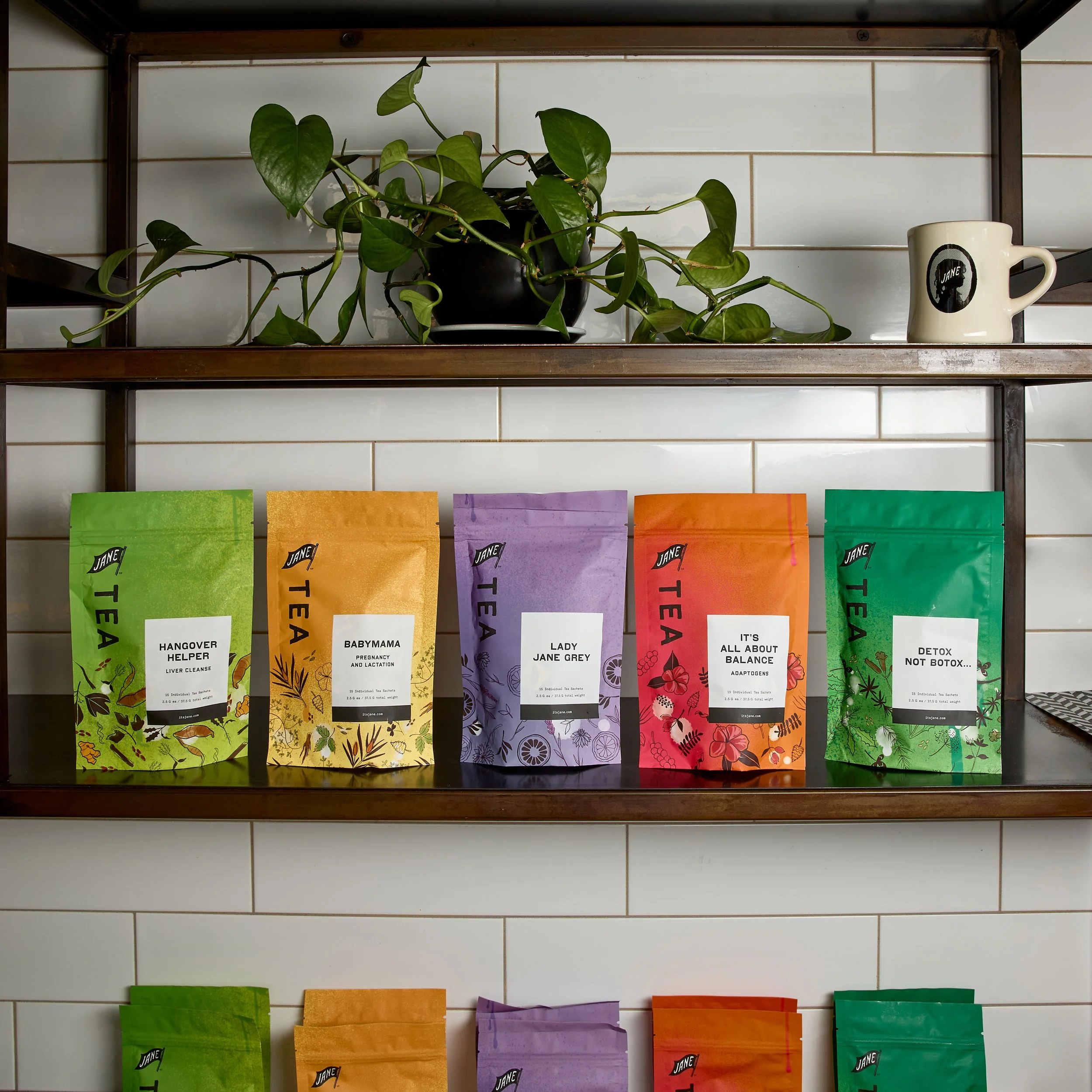 A wooden shelf with a green potted plant and a white mug on top, and five colorful packages of herbal tea on the shelf below, against a tiled wall.