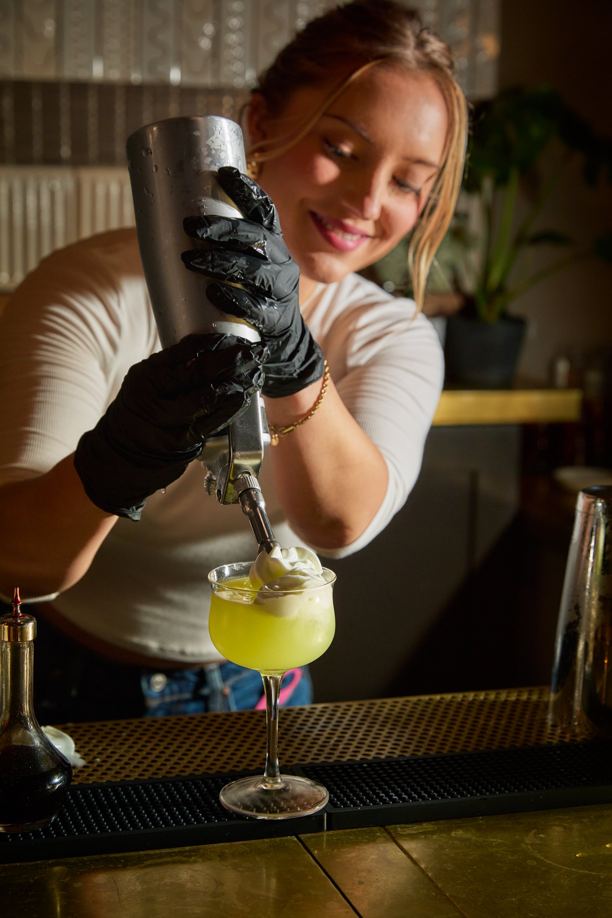 A woman wearing black gloves pours whipped cream into a yellow-green cocktail drink at a bar.