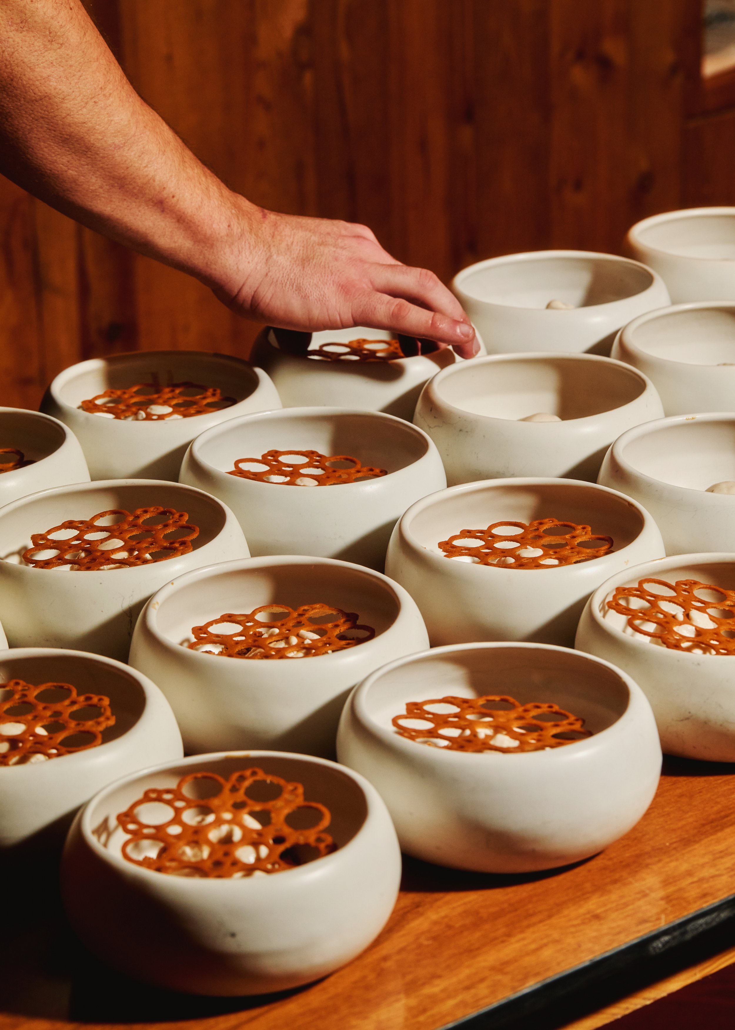 A person’s hand reaching over multiple white ceramic bowls filled with stew, each topped with pretzel sticks, on a wooden table.