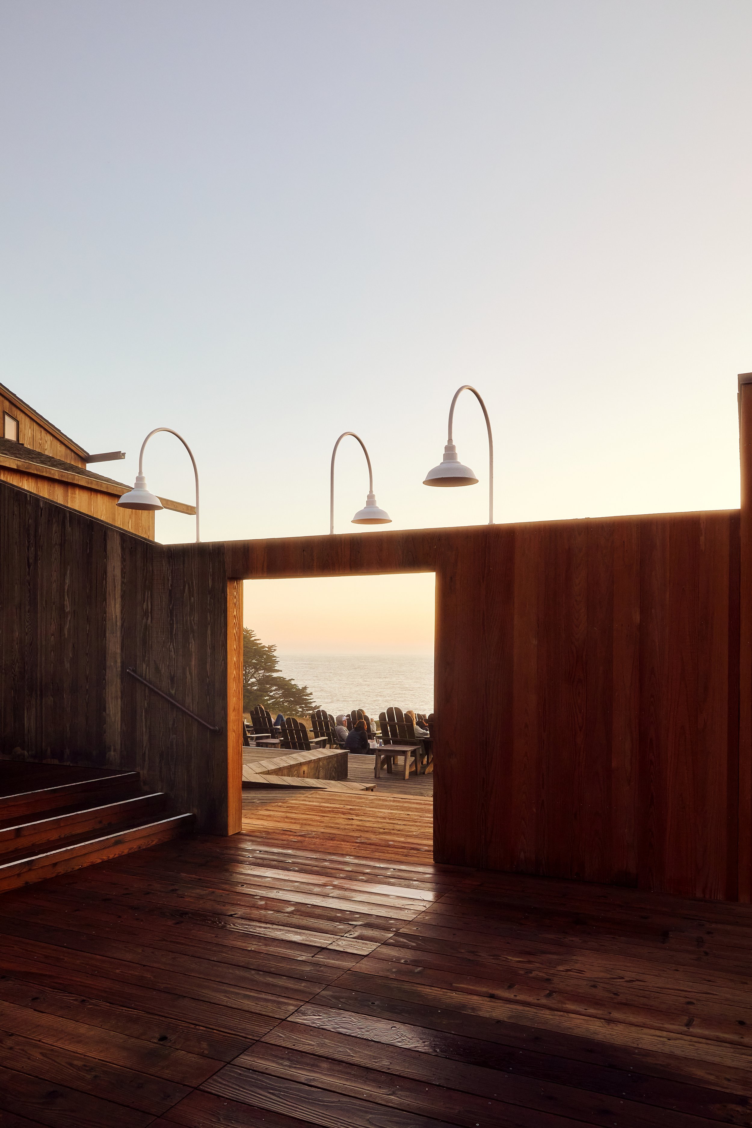 View of a wooden outdoor deck at sunset with ocean in the background, overlooking Adirondack chairs and people.