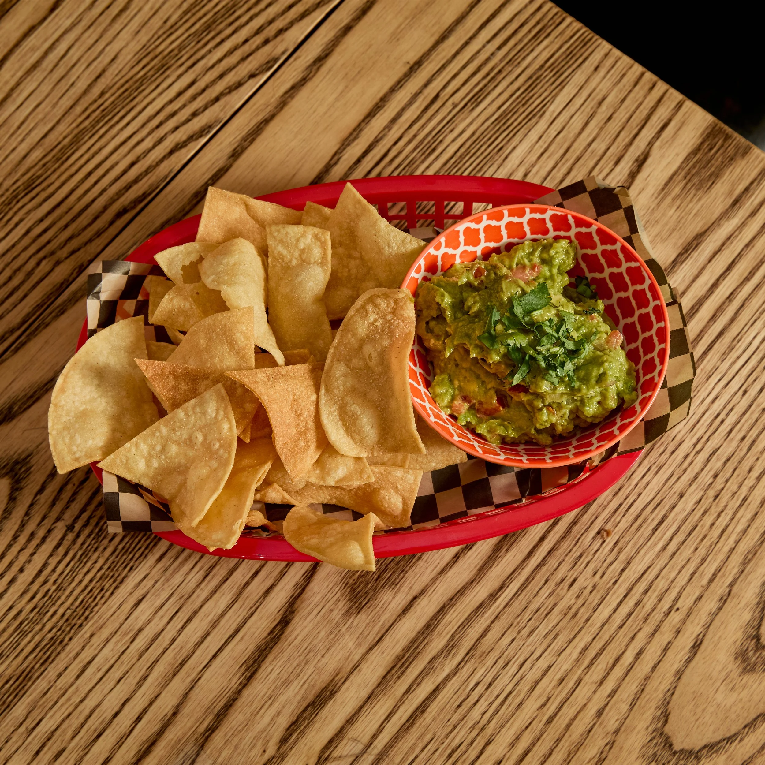 A basket of tortilla chips with guacamole in a patterned bowl on a wooden table.