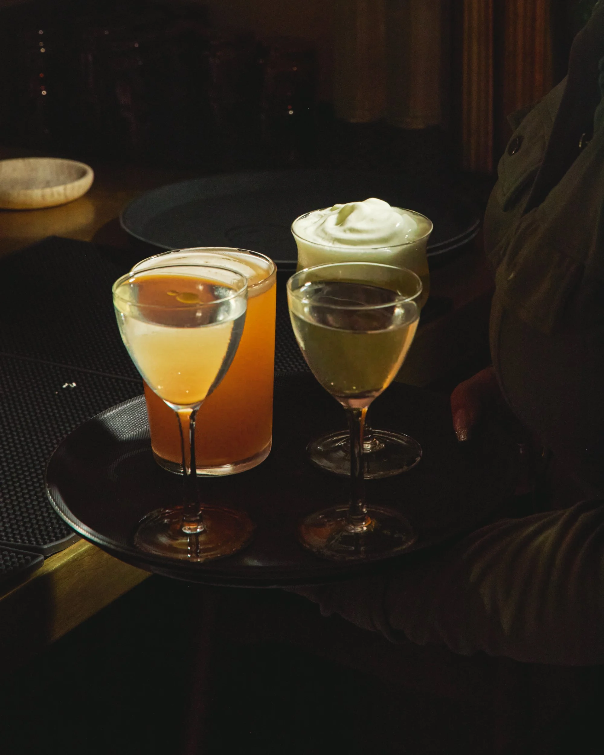 A person holding a black tray with three cocktails: two in wine glasses and one in a highball glass, all garnished with whipped cream, on a dark table in a dimly lit setting.