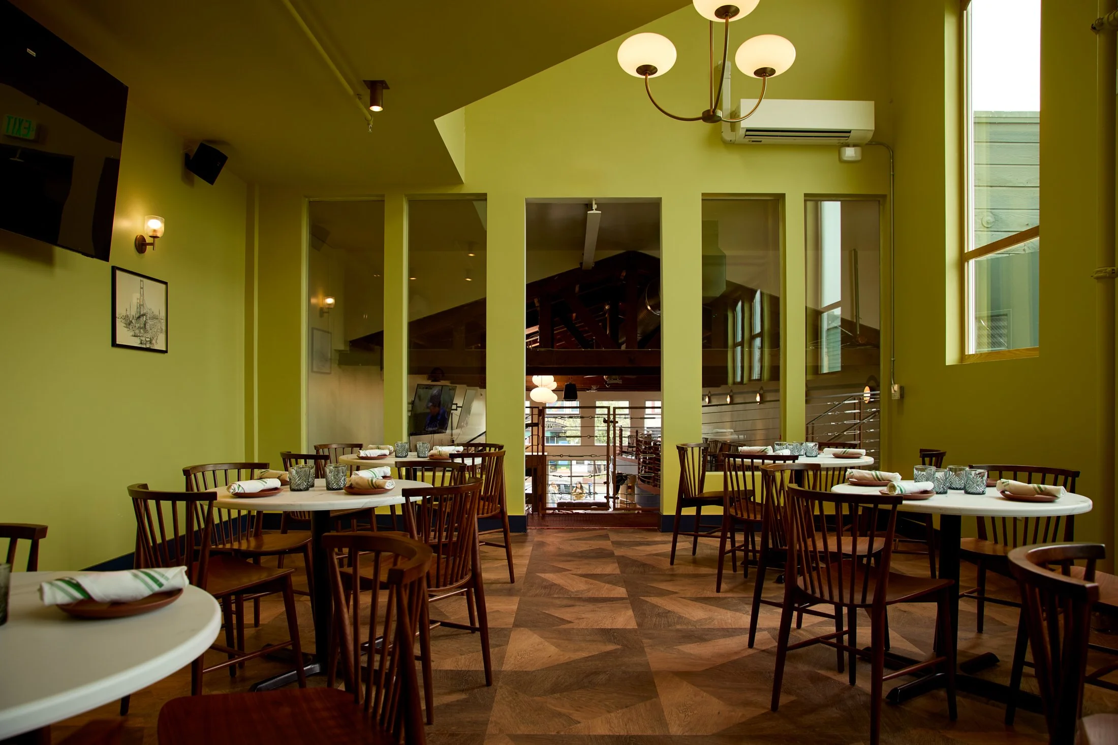 Interior of a restaurant with lime green walls, multiple round tables set with napkins and drinking glasses, wooden chairs, large windows, a chandelier, a wall-mounted TV, and a visible staircase in the background.