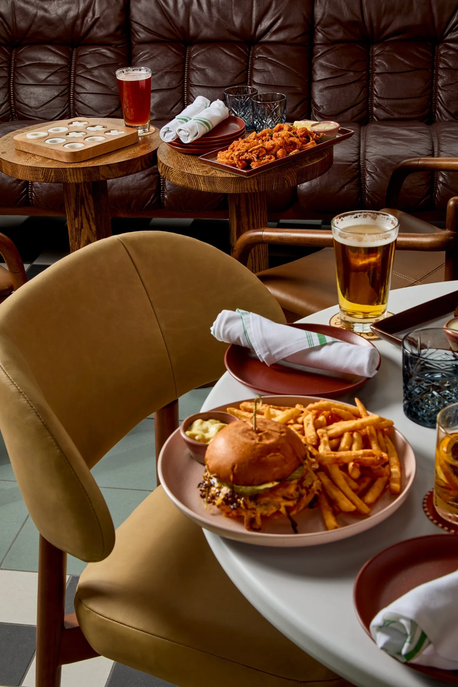 Table with a plate of fried chicken, fries, a cheeseburger, small dishes of dipping sauces, and drinks in glasses. Background includes cushioned seating and another table with similar setup.