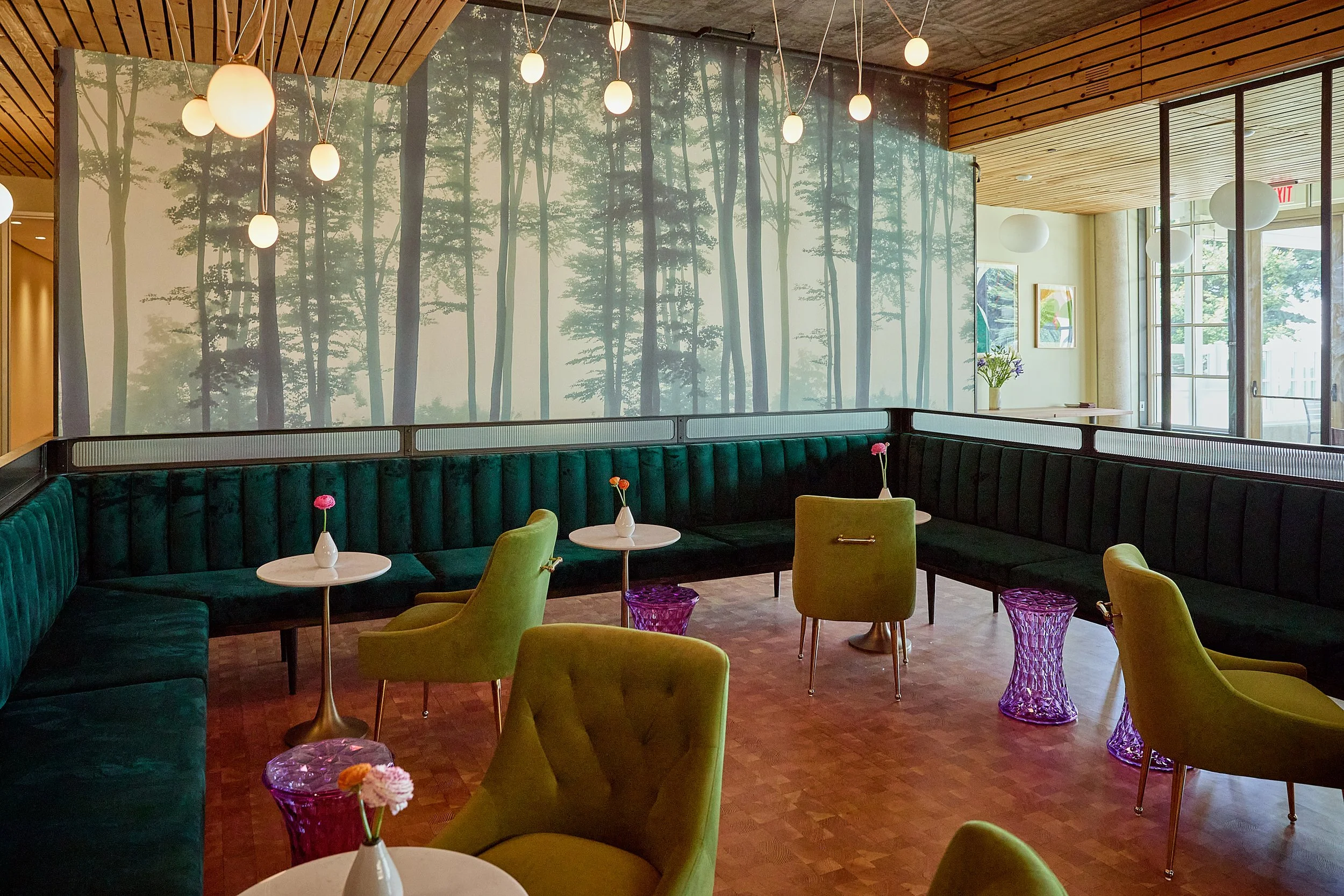 Interior of a modern restaurant with a large forest-themed mural, hanging light fixtures, green velvet seating, and glass-topped tables with small flower arrangements.