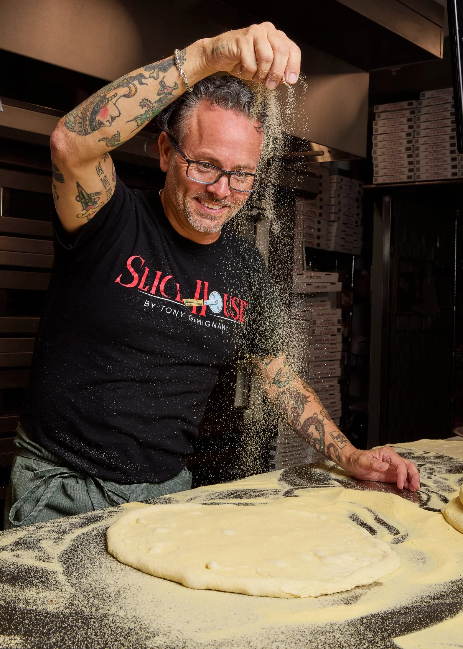 A man with tattoos and glasses is sprinkling flour on a pizza dough that is rolled out on a floured surface, in what appears to be a pizzeria kitchen.