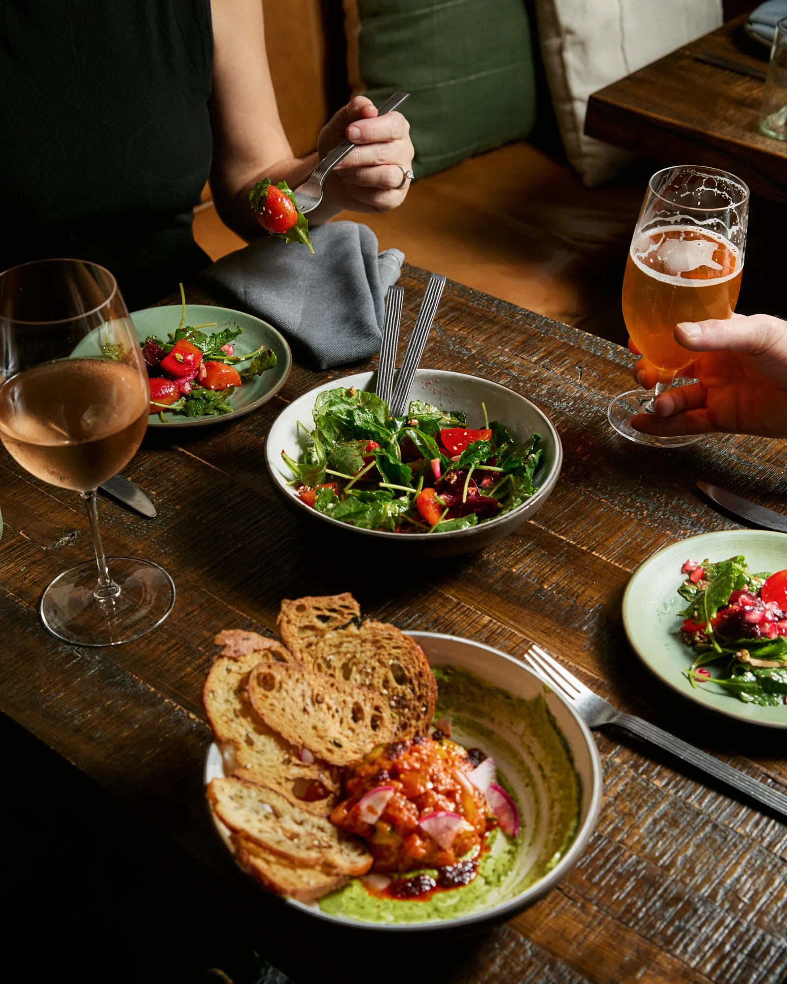 People sharing a meal at a wooden table, with salads, bread, and beverages, including a glass of rosé wine and a glass of beer.