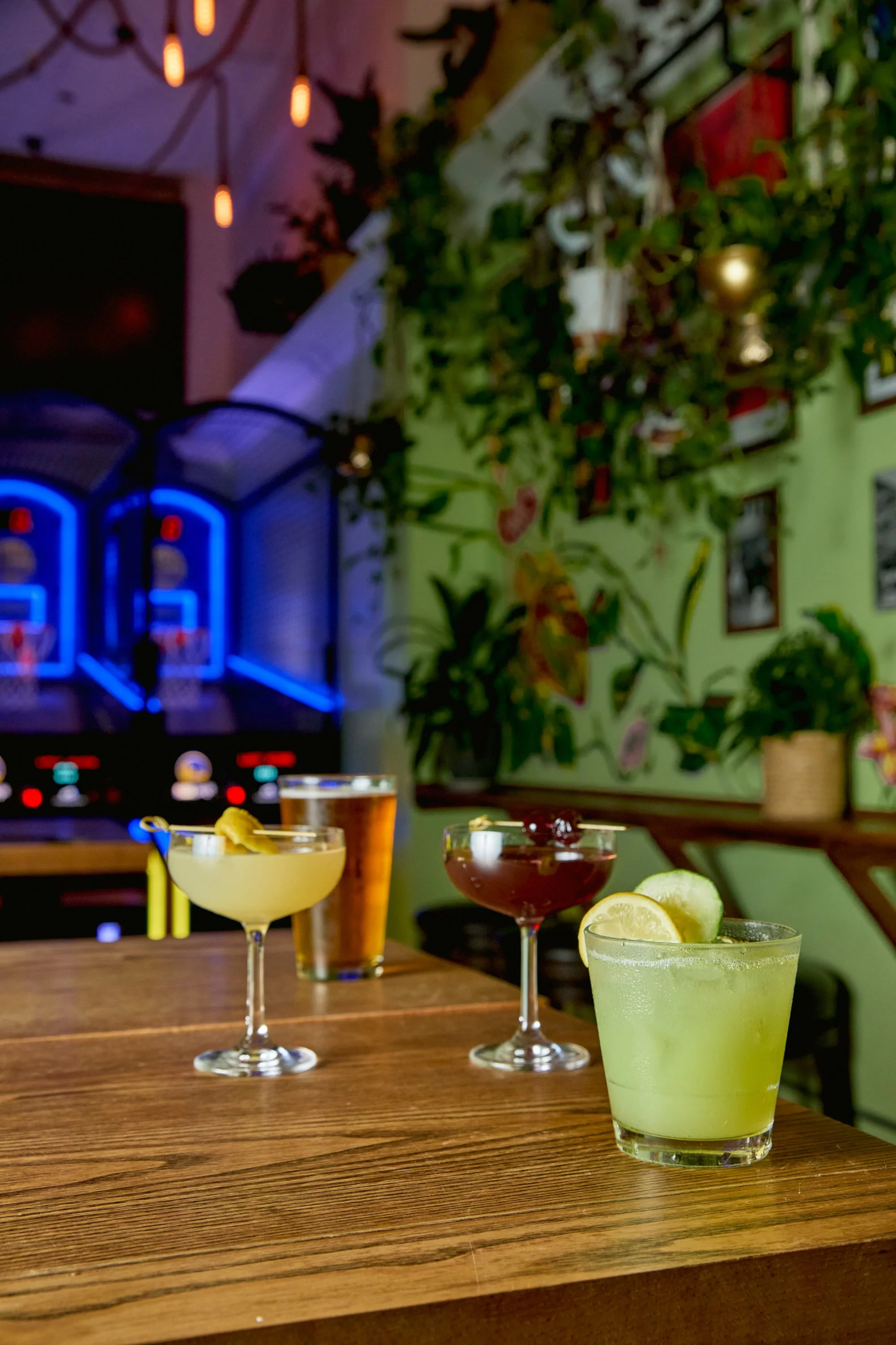 Four cocktails on a wooden table inside a bar or restaurant, with a green wall decorated with plants in the background and electronic dartboards on the wall.
