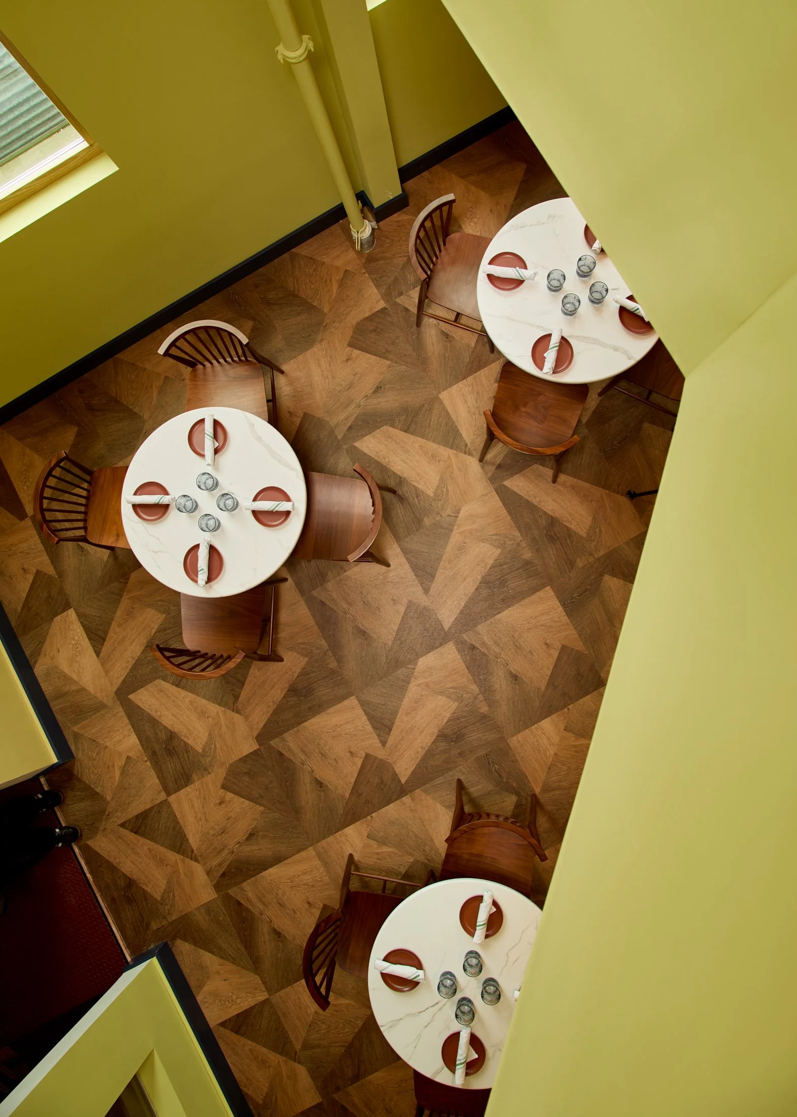 Two round white tables set for dining with four glasses and four red plates each, four napkins placed on each plate, placed in a room with wooden parquet floors, yellow walls, and a window with blinds, viewed from above.