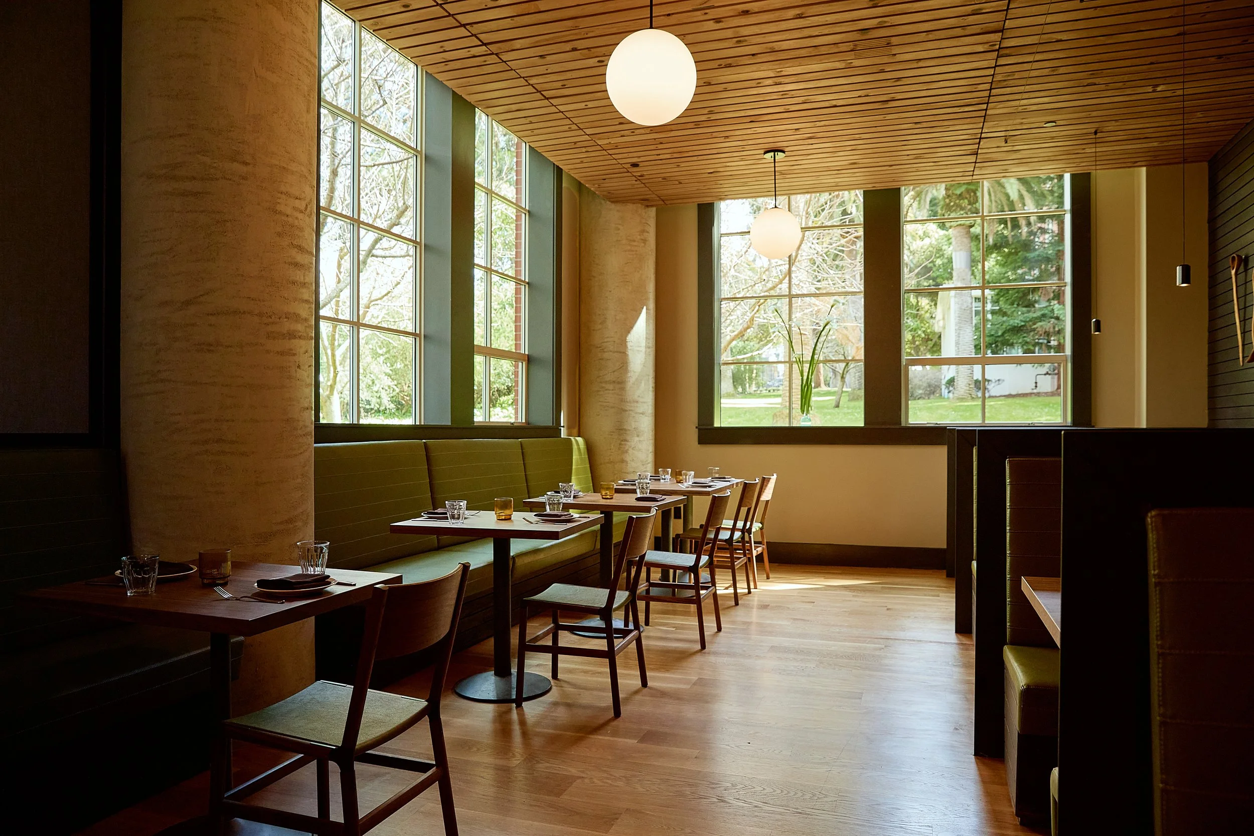 Interior of a modern restaurant with large windows, wooden ceiling, and minimal decor. Tables are set with glasses, plates, and utensils, and sunlight streams through the windows, revealing a view of trees outside.