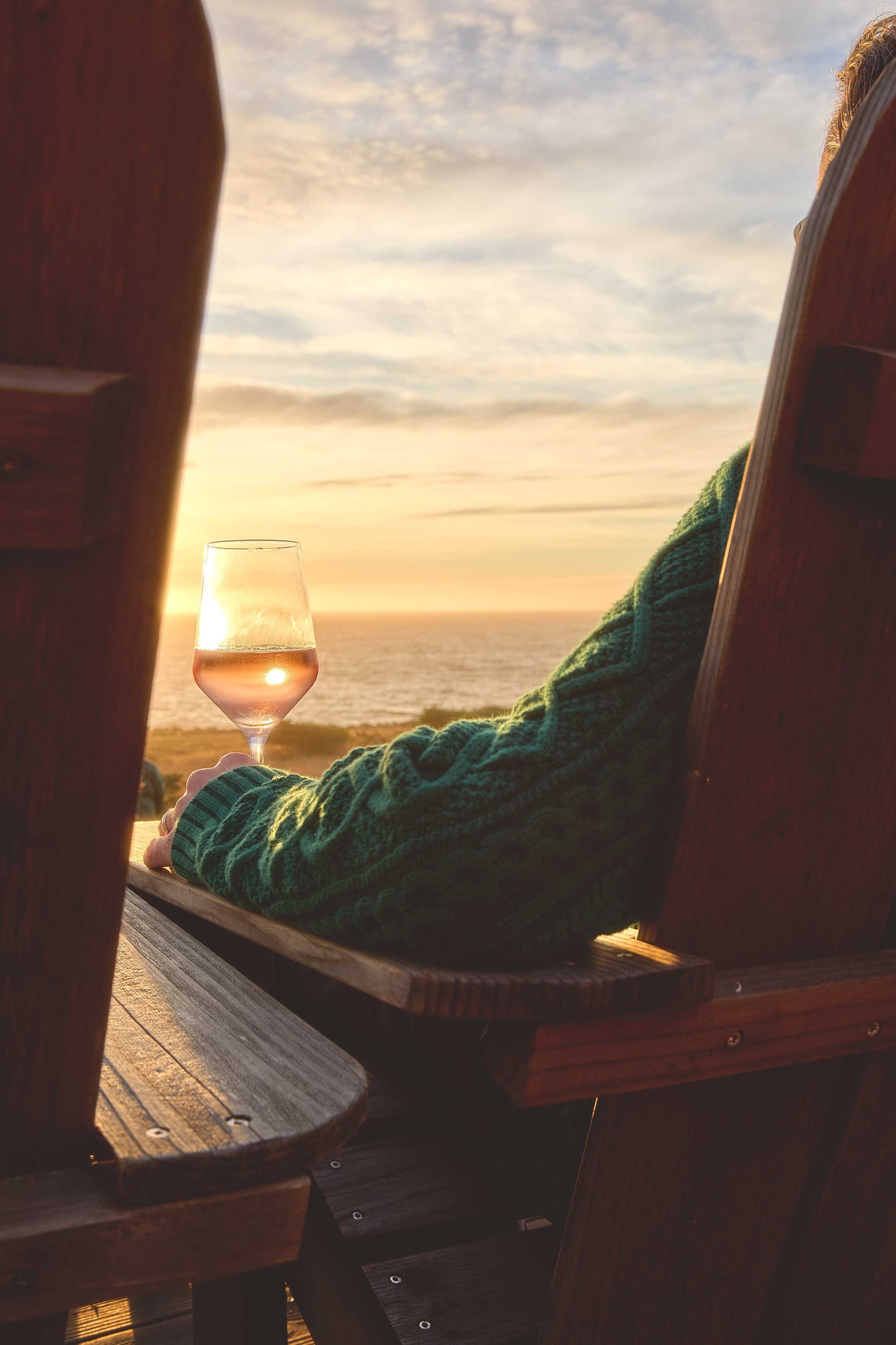 Person in a green sweater sitting outdoors with a glass of rosé wine, watching a sunset over the ocean, framed by wooden chairs.