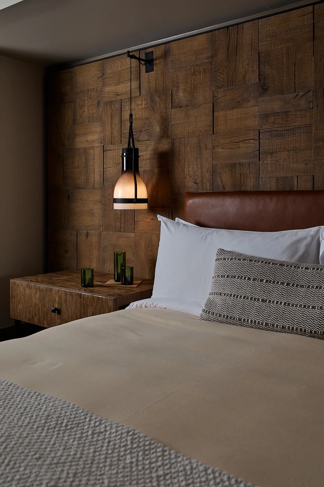 A cozy bedroom featuring a bed with white and patterned pillows, a wooden headboard, a wooden nightstand with three green glass vases, and a modern hanging lamp with a warm glow.