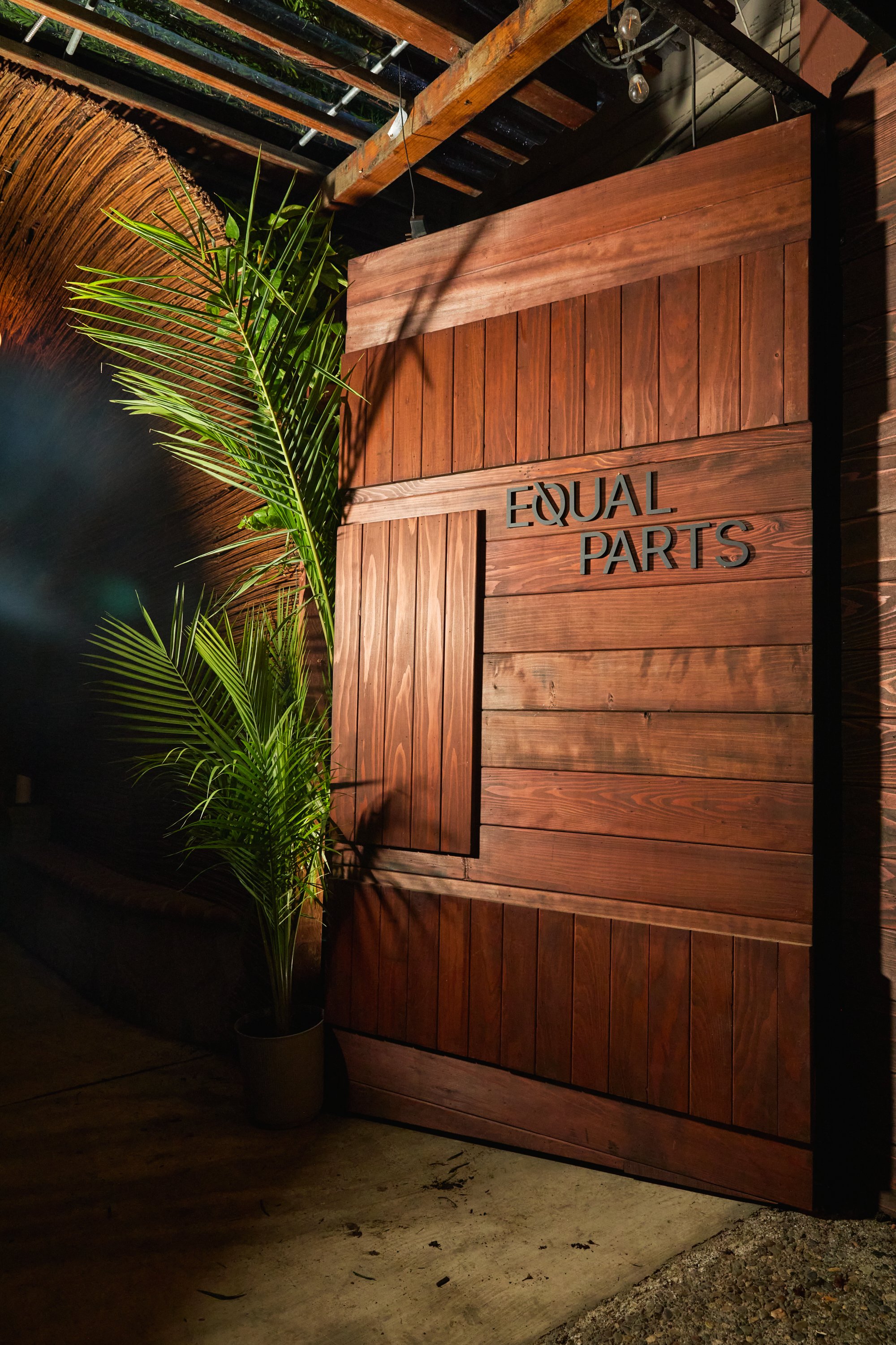 A wooden wall with a sign reading "EQUAL PARTS" next to a potted plant with large green leaves, set against a dimly lit background with exposed wooden beams and hanging light bulbs.