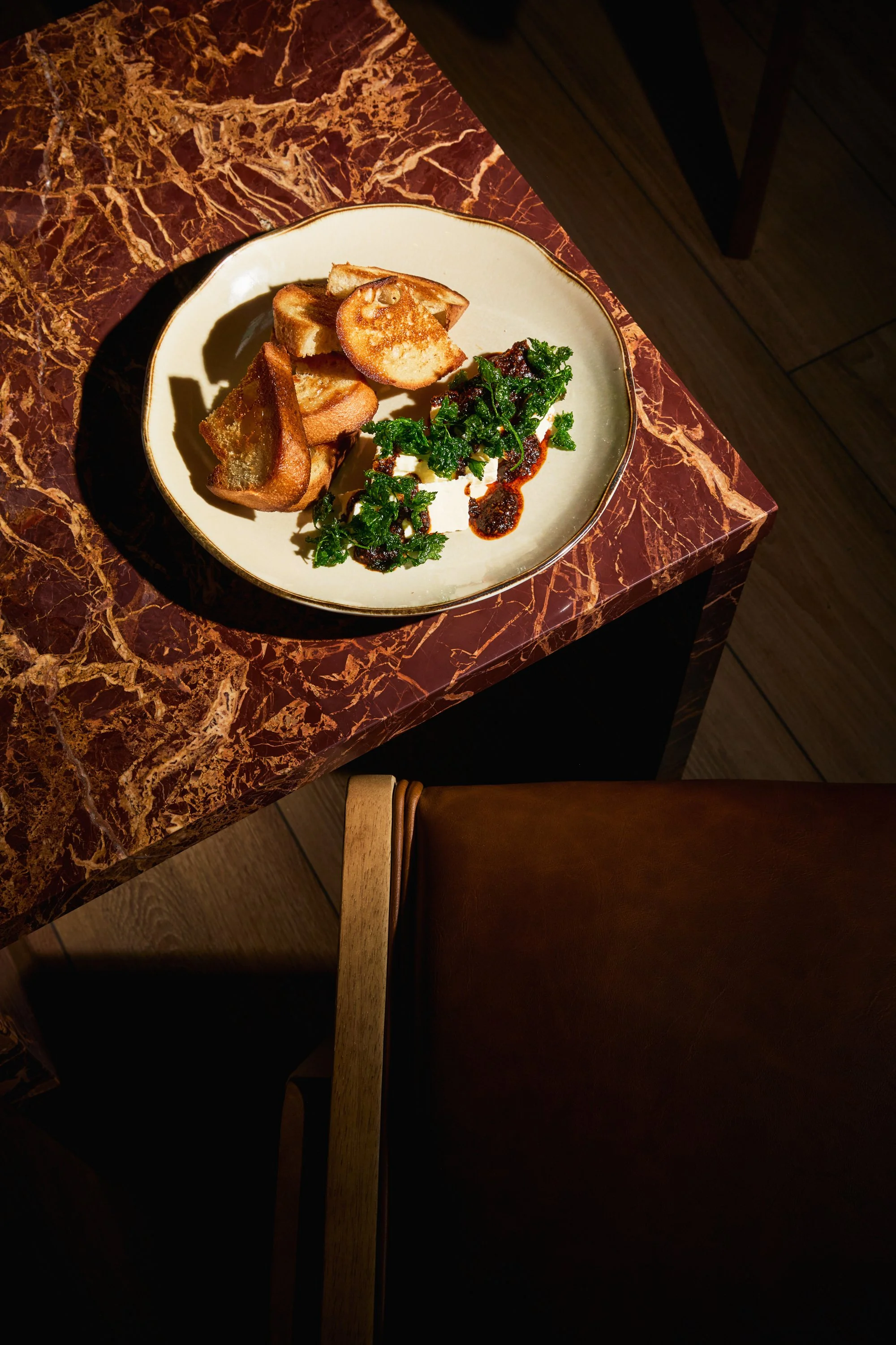 A plate with toasted bread, greens, and sauce on a marble table with a wooden chair nearby.