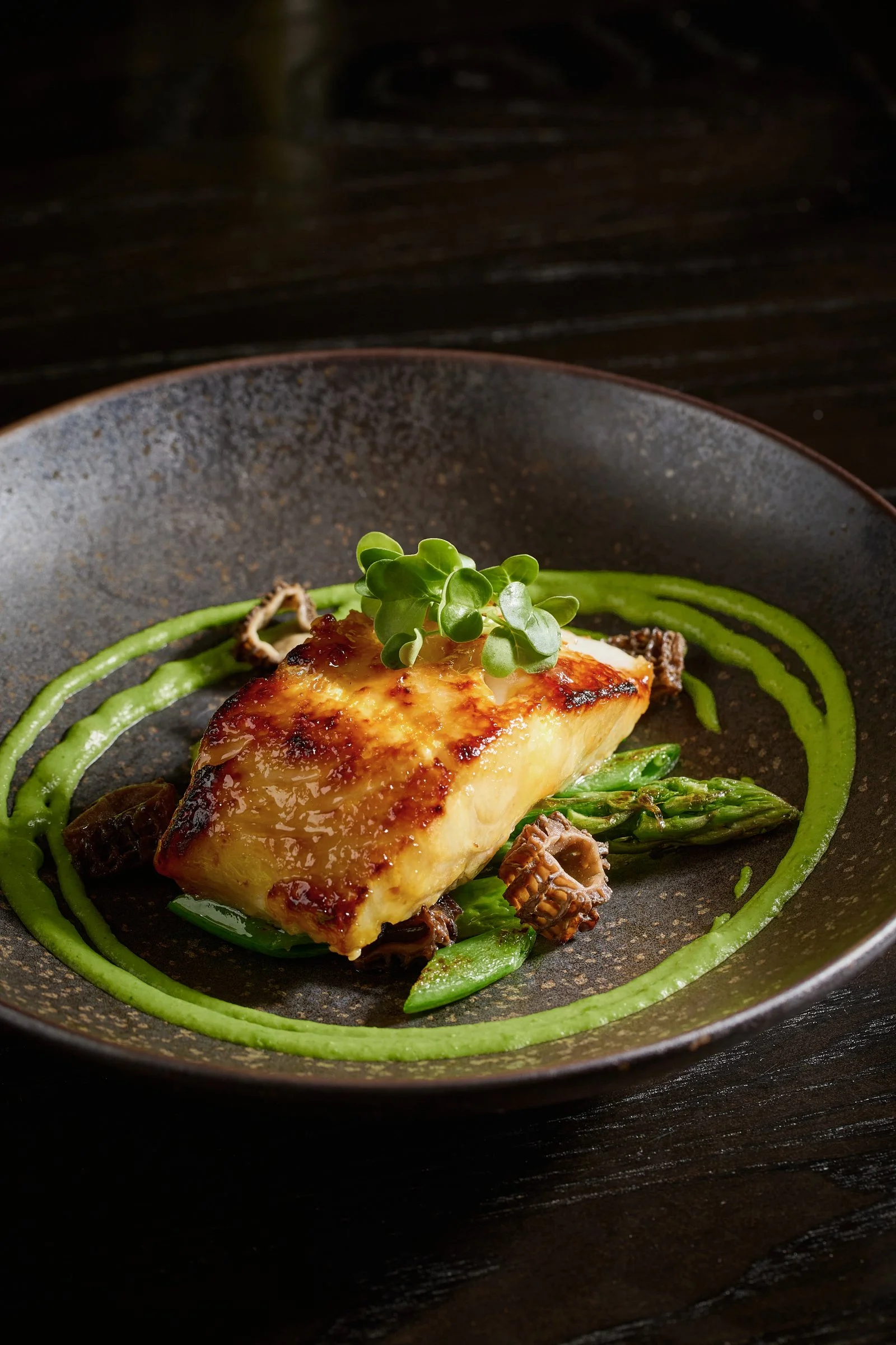 A plated dish with a piece of seared fish, garnished with microgreens, served on a bed of green vegetables, and surrounded by a green sauce spiral.