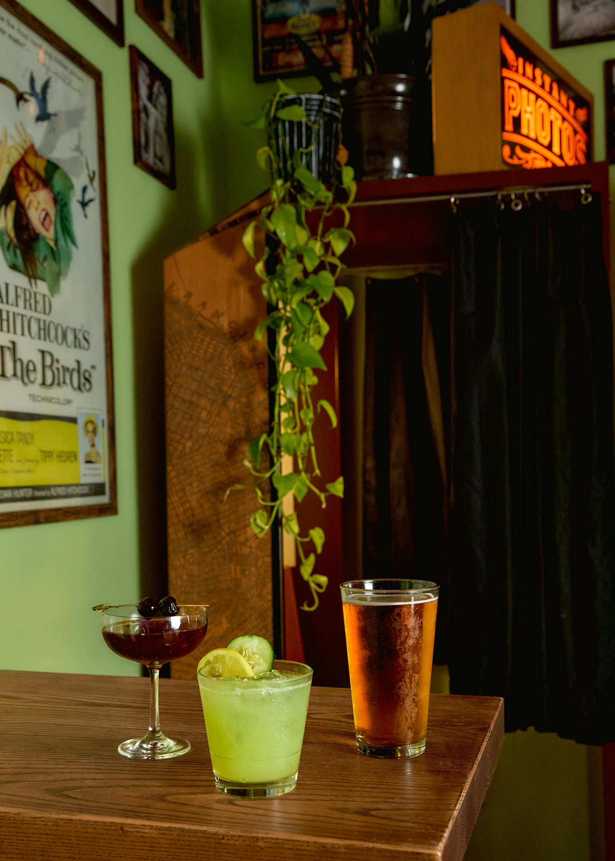 Three cocktails on a wooden table: a dark red drink with cherries, a green drink with cucumber and lemon slices, and an amber-colored beer. The background shows a green wall with framed posters, a potted plant hanging, and a vintage photo booth sign.