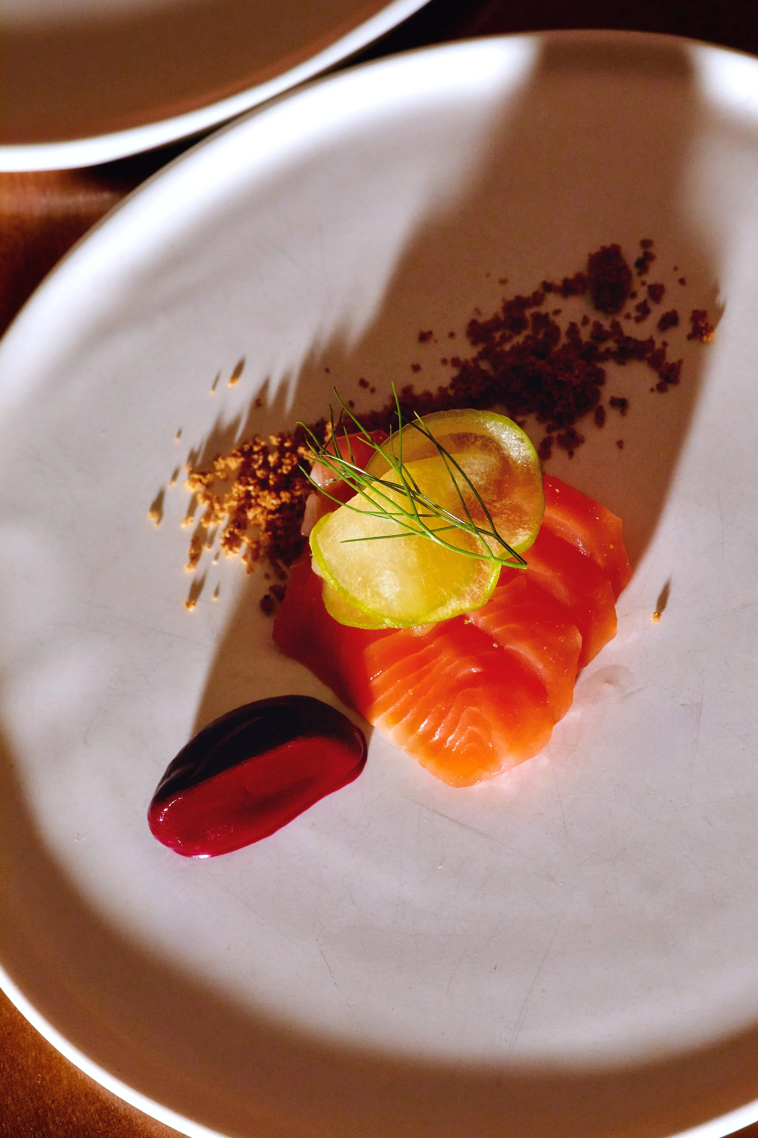 A plated dish with slices of salmon, a grape tomato, a slice of cucumber, a dollop of red sauce, and some garnishes on a white plate.