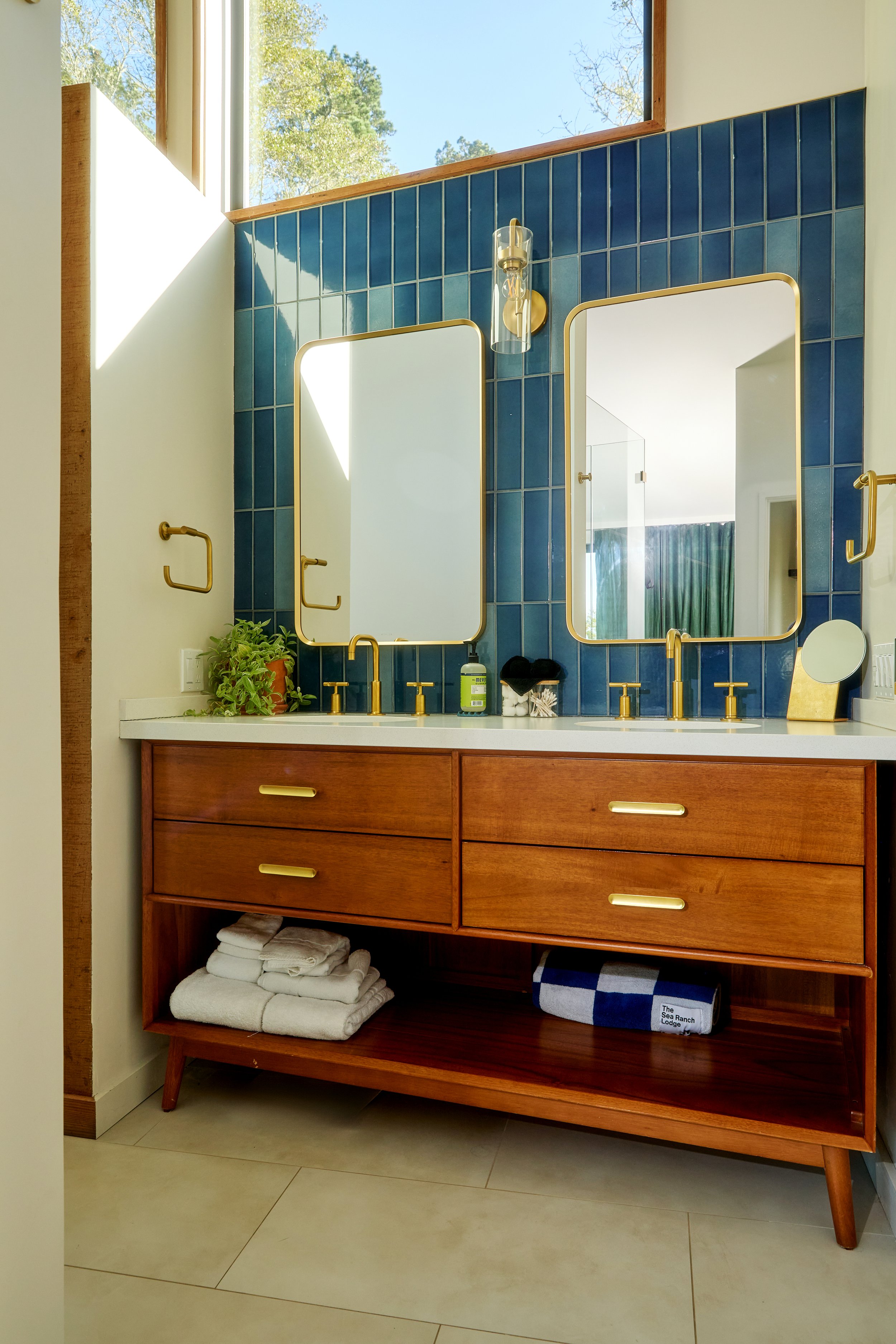 Modern bathroom vanity with a white countertop, gold fixtures, and two mirrors framed in gold, set against a blue tiled wall with a small plant and toiletries on the counter, and towels stored on the lower shelf.