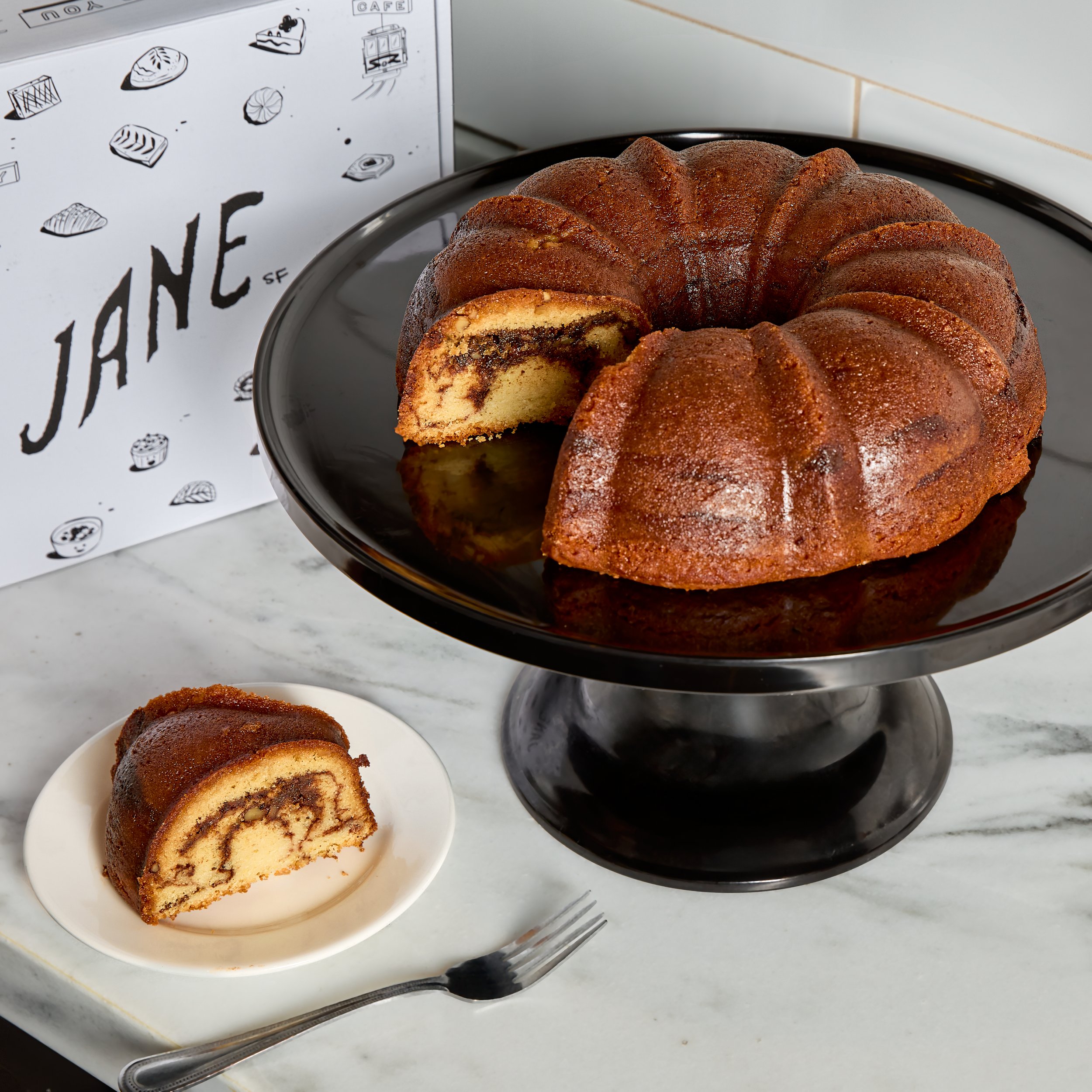 A marble Bundt cake with a slice cut out and placed on a white plate. The cake has a chocolate swirl inside and is on a black cake stand, with a fork next to the plate. There is a sign in the background with the word "JANE" and small sketches of bake