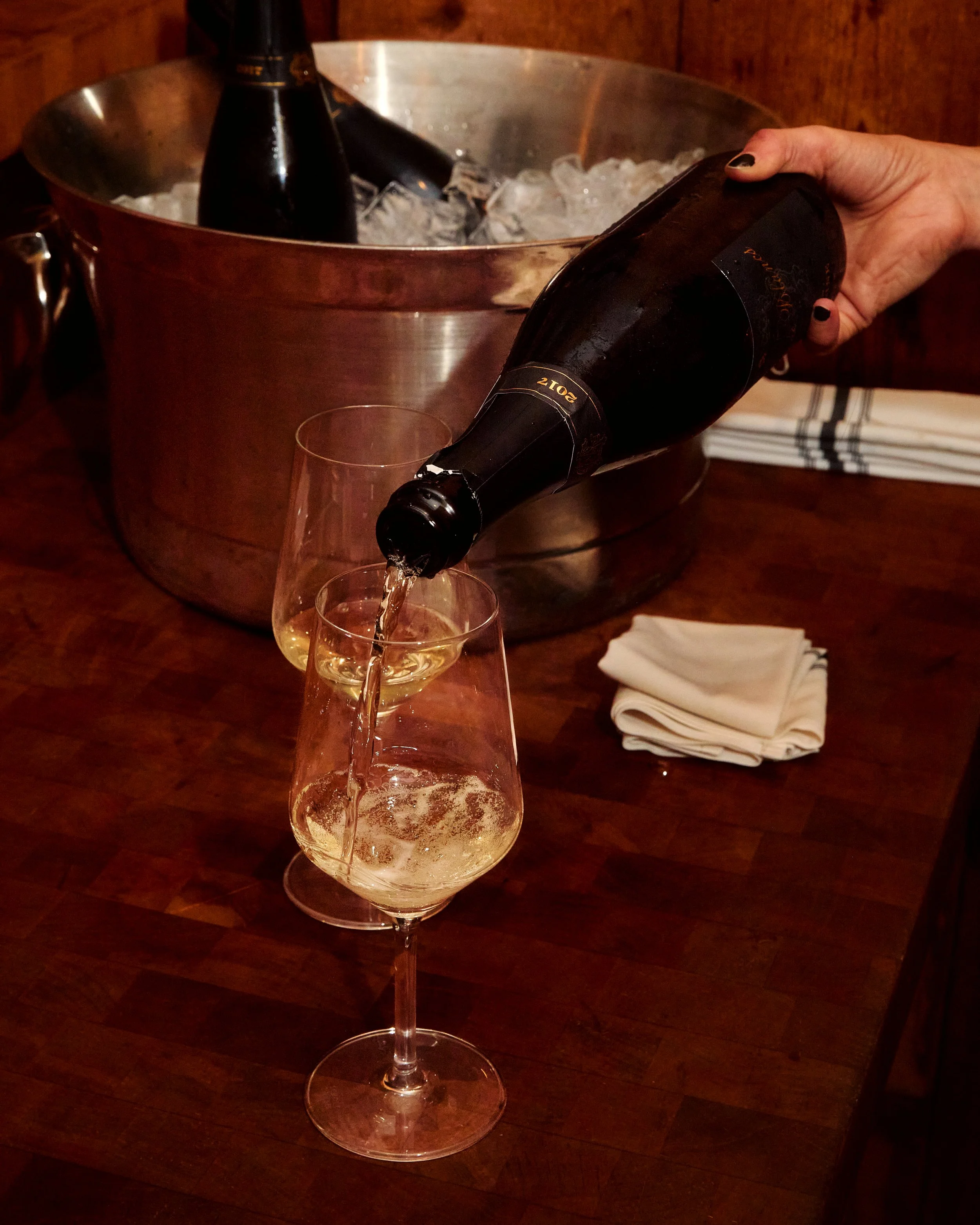 Person pouring white wine into a champagne flute, with a bucket of ice and bottles of champagne in the background.