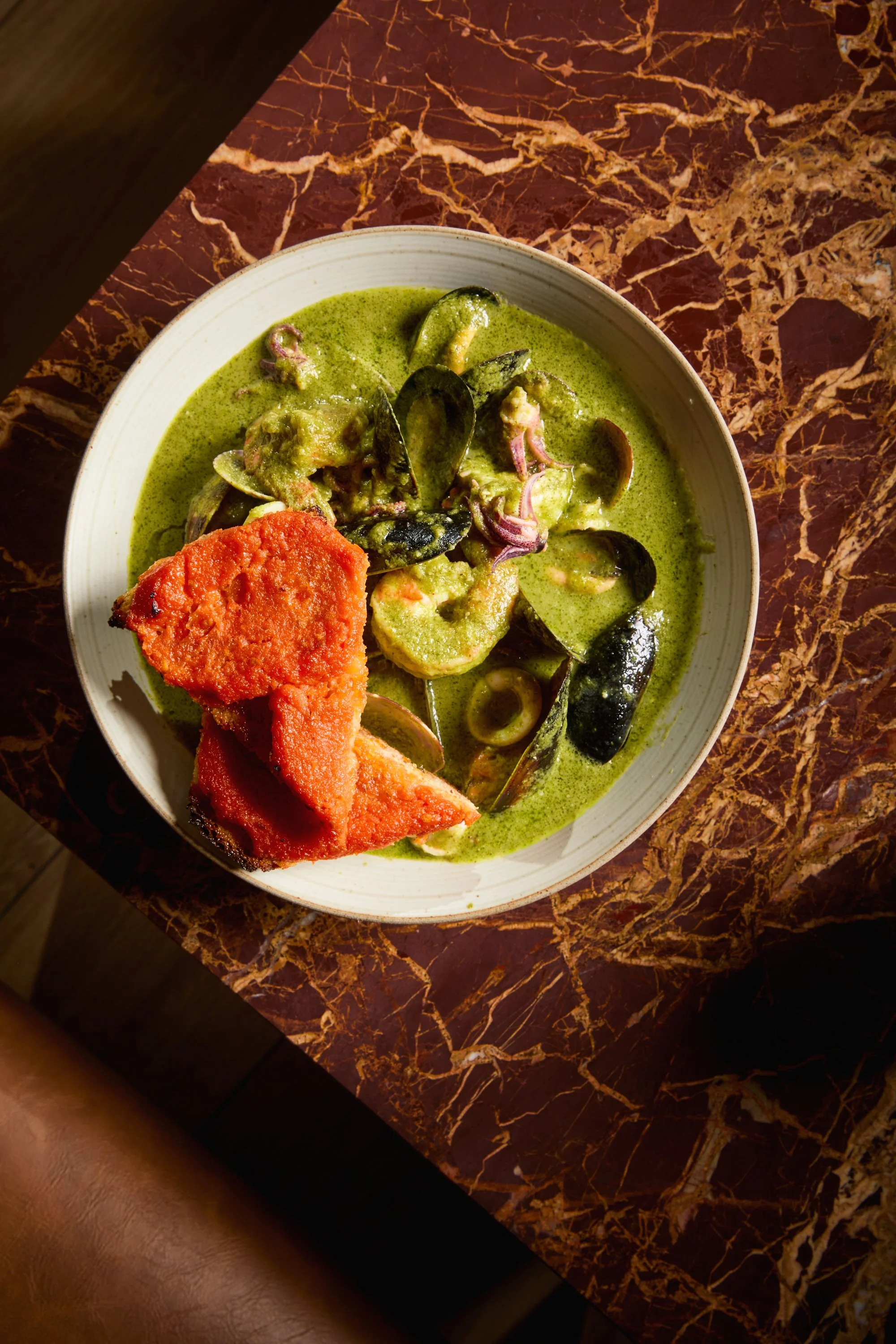A bowl of green curry with vegetables and fried bread on a dark marble table.