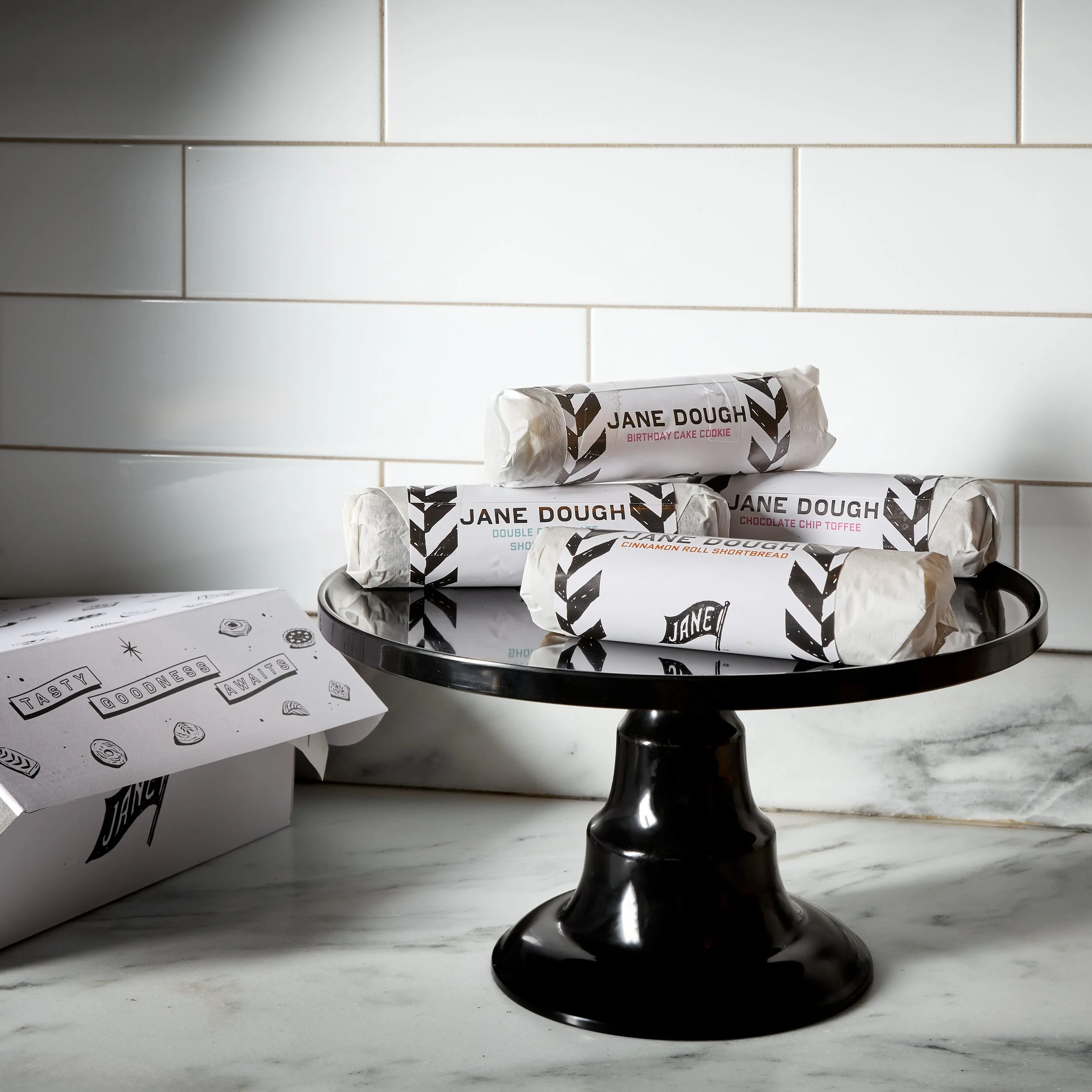 Wrapped cookies from Jane Dough on a black cake stand in a white tiled kitchen with a white box labeled 'TASTE. GOOD. WAIT.'