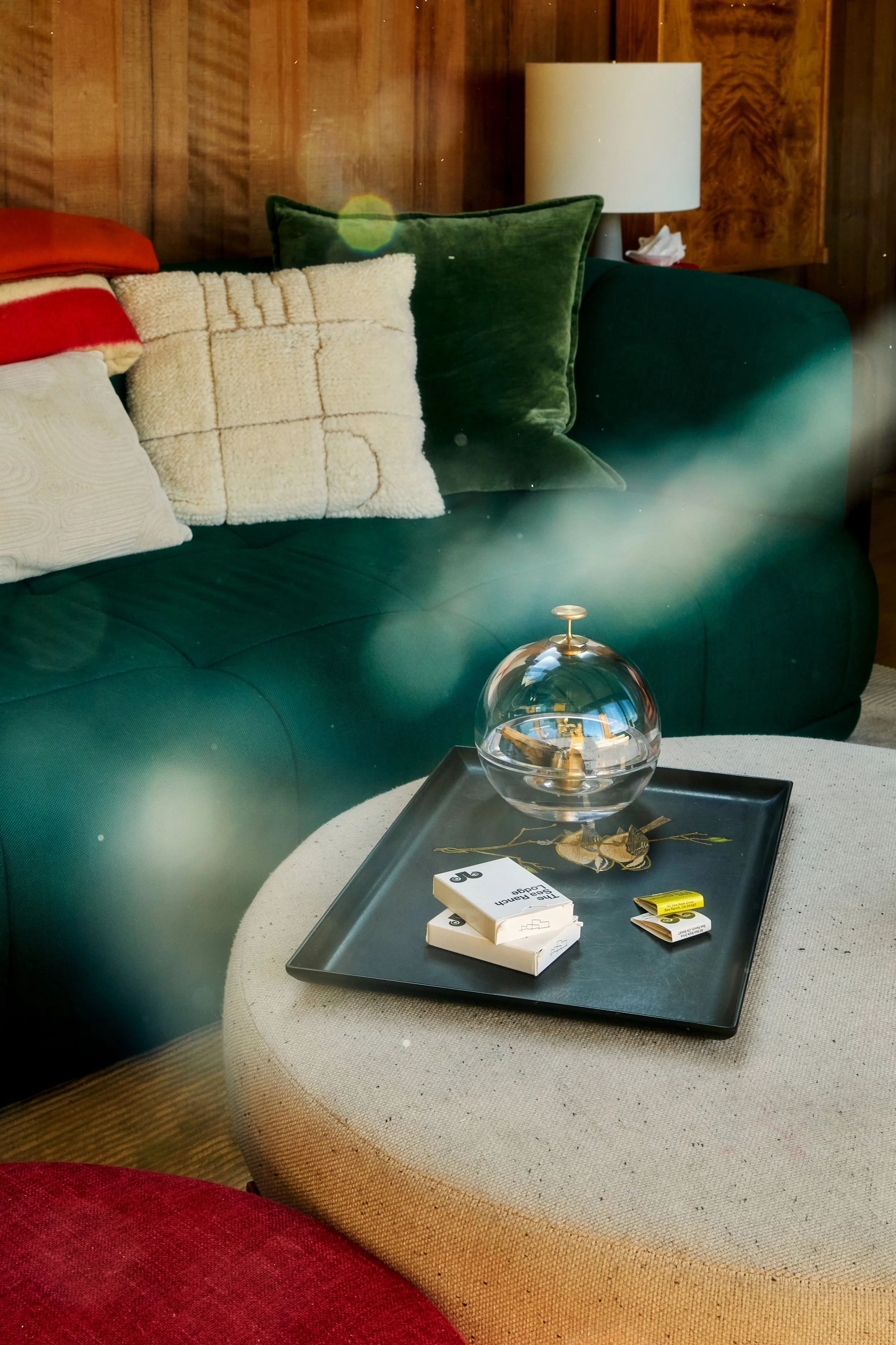 Living room sofa with assorted pillows, a coffee table with a decorative glass orb and matches, in front of wooden paneled wall and side lamp.