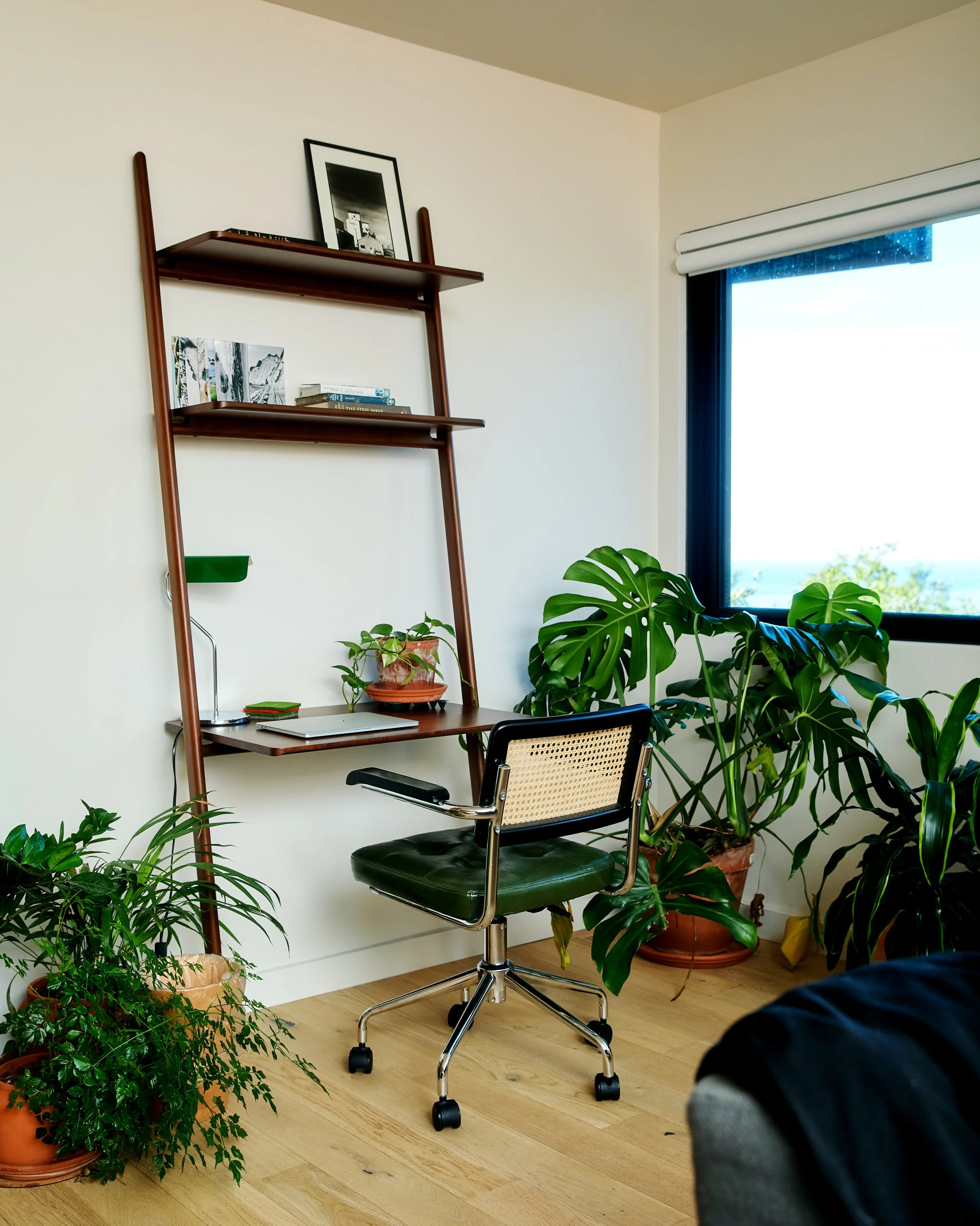 A home office corner with a vintage green office chair, plants on the floor and windowsill, a wooden ladder-style bookshelf with books and framed art, and a window with a view of greenery outside.